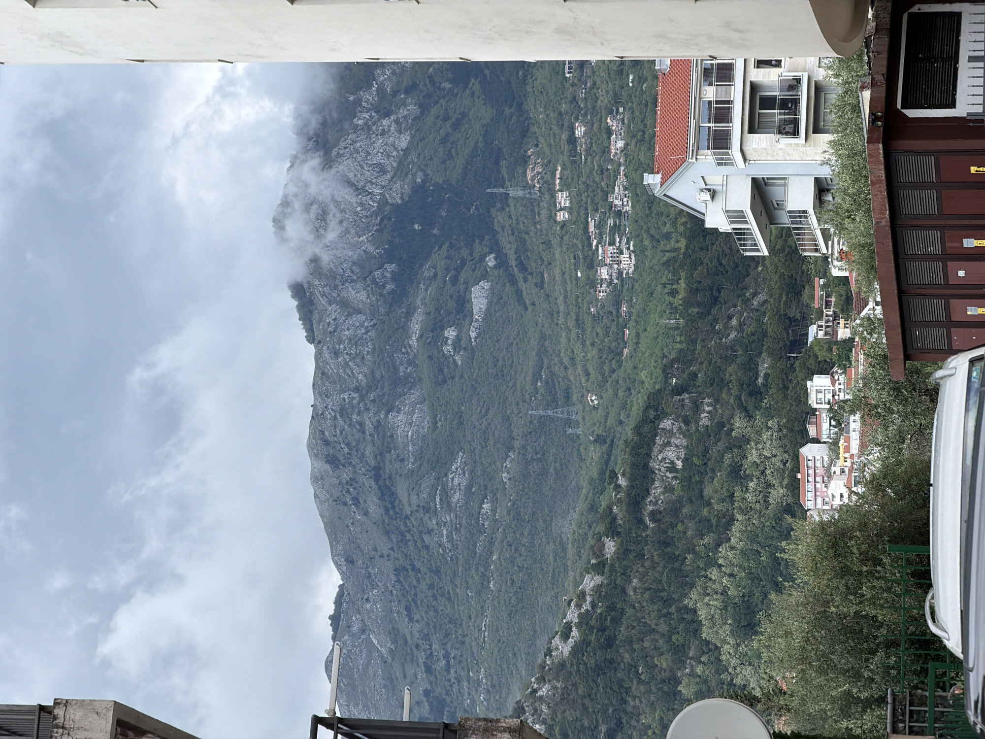 The image depicts a view from a building in Budva, Montenegro, looking towards a large, verdant mountain partially obscured by clouds. The foreground includes elements of the building from which the photo was taken, such as a roof, railing, and a satellite dish. Adjacent to the viewpoint is another building with red-tiled roofs and a multi-story structure. The mountain slopes are covered in dense vegetation, interspersed with occasional houses and power lines. Buildings are nestled within the lush greenery, creating a sense of habitation integrated into the natural landscape. A white van is partly visible on the right edge of the image. The sky is overcast, with clouds enveloping the upper part of the mountain, obscuring its peak.