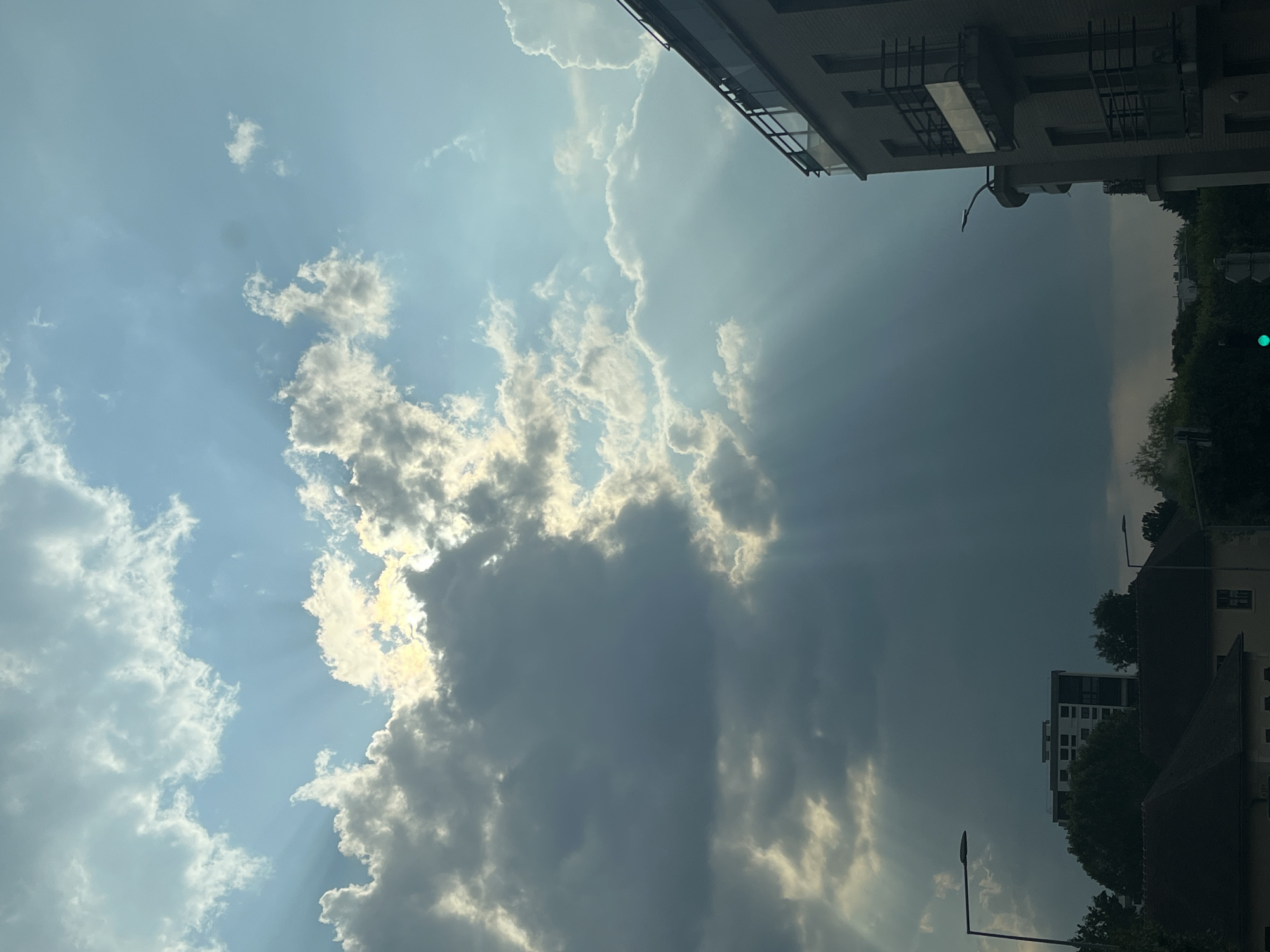 The image shows a sky with dramatic cloud formations and sunbeams, characteristic of a partly cloudy day, possibly with approaching rain. In the foreground, a multi-story building with balconies is visible on the right side. In the distance, on the left, are other buildings, including a taller, light-colored apartment building, and residential rooftops. Trees and streetlights are also present in the lower portions of the frame. The scene appears to be urban or suburban. Based on the context provided, this scene is located in the city of L'Haÿ-les-Roses, France. There are no people or animals clearly visible in the image. The overall lighting suggests it is daytime.