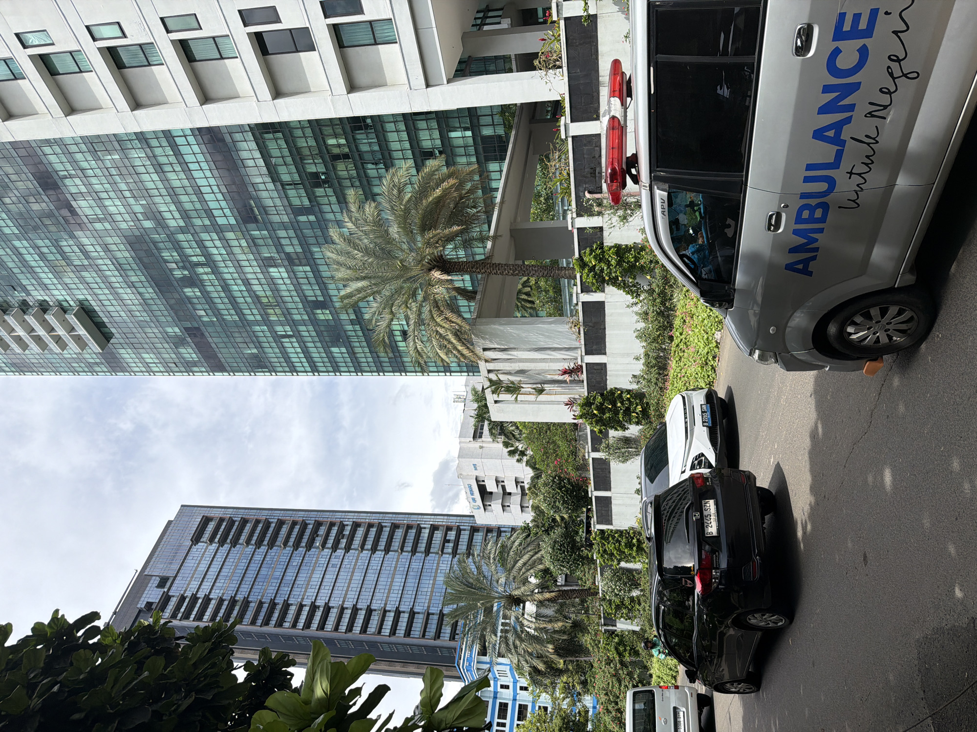 The image shows a street scene in South Jakarta, Indonesia. Tall buildings with glass facades are visible on the left side of the frame. Palm trees are situated along the street. Several vehicles are parked on the right side, including a silver ambulance with the word "AMBULANCE" in blue letters, along with "Untuk Negeri." A black SUV and a white car are also parked. The sky is overcast.