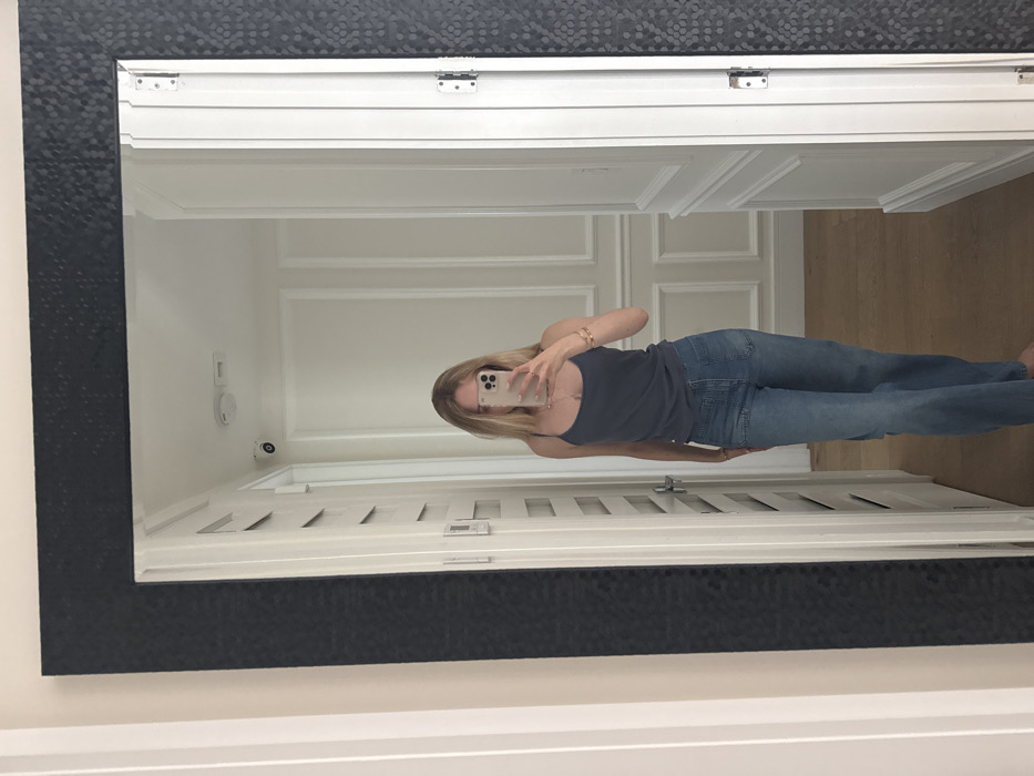 An indoor scene in Coral Gables, United States of America, depicts a full-body mirror selfie. A female subject with long blonde hair, dressed in a dark blue spaghetti-strap tank top and light blue wide-leg jeans, is captured in the reflection of a large mirror. Her face is obscured by a light-colored smartphone she holds with both hands, actively taking the photograph. She wears a necklace with a pendant and multiple bracelets on her left wrist. The mirror features a wide, dark-colored, textured frame.
The background, visible within the mirror's reflection, shows a well-lit interior space with white paneled walls adorned with decorative molding and light wooden flooring. A white dome-shaped security camera and a white smoke detector are mounted on the white ceiling. On the left side of the reflection, white louvered bifold doors are visible. A standard white door is seen on the right, featuring two silver hinges on its edge, indicating it is a functioning door or the mirror is positioned next to one.