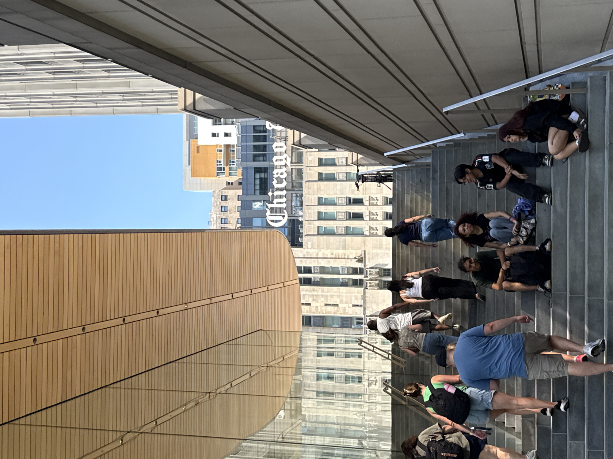 The image depicts an outdoor scene with a group of people on a wide, modern stone staircase. The staircase is flanked by a building with a stone facade and a large, cantilevered wooden overhang with glass walls. In the background, several buildings are visible, including one with a prominent "Chicago" sign. The sky is clear and blue.
Several individuals are present on the stairs. Some are walking up the stairs, while others are sitting or resting on the steps.
On the lower part of the stairs, a man in a blue t-shirt and khaki shorts is walking upwards. To his left, a woman in a green tank top and denim shorts with a black backpack is ascending. Further to the left, a person with a backpack and shorts is also moving up.
On the middle section of the stairs, two individuals are sitting. One is a person in a dark t-shirt and jeans, and the other is a young woman in a black top and jeans. To their right, another person in a black t-shirt and black pants is sitting.
Further up the stairs, a woman in a white tank top and black pants is walking upwards, followed by another woman in a blue shirt and jeans. Ahead of them, a person in a white tank top and shorts is also ascending. On the upper part of the stairs, a person in a camouflage-patterned shirt and jeans is walking up, and another person in a white tank top and dark shorts is also ascending.
A black bicycle is parked upright on the stairs in the mid-ground. The architectural style suggests a modern urban setting, likely a public space or an entrance to a large building in Chicago, USA, given the "Chicago" signage. The overall atmosphere is casual and leisurely, with people navigating the public space.