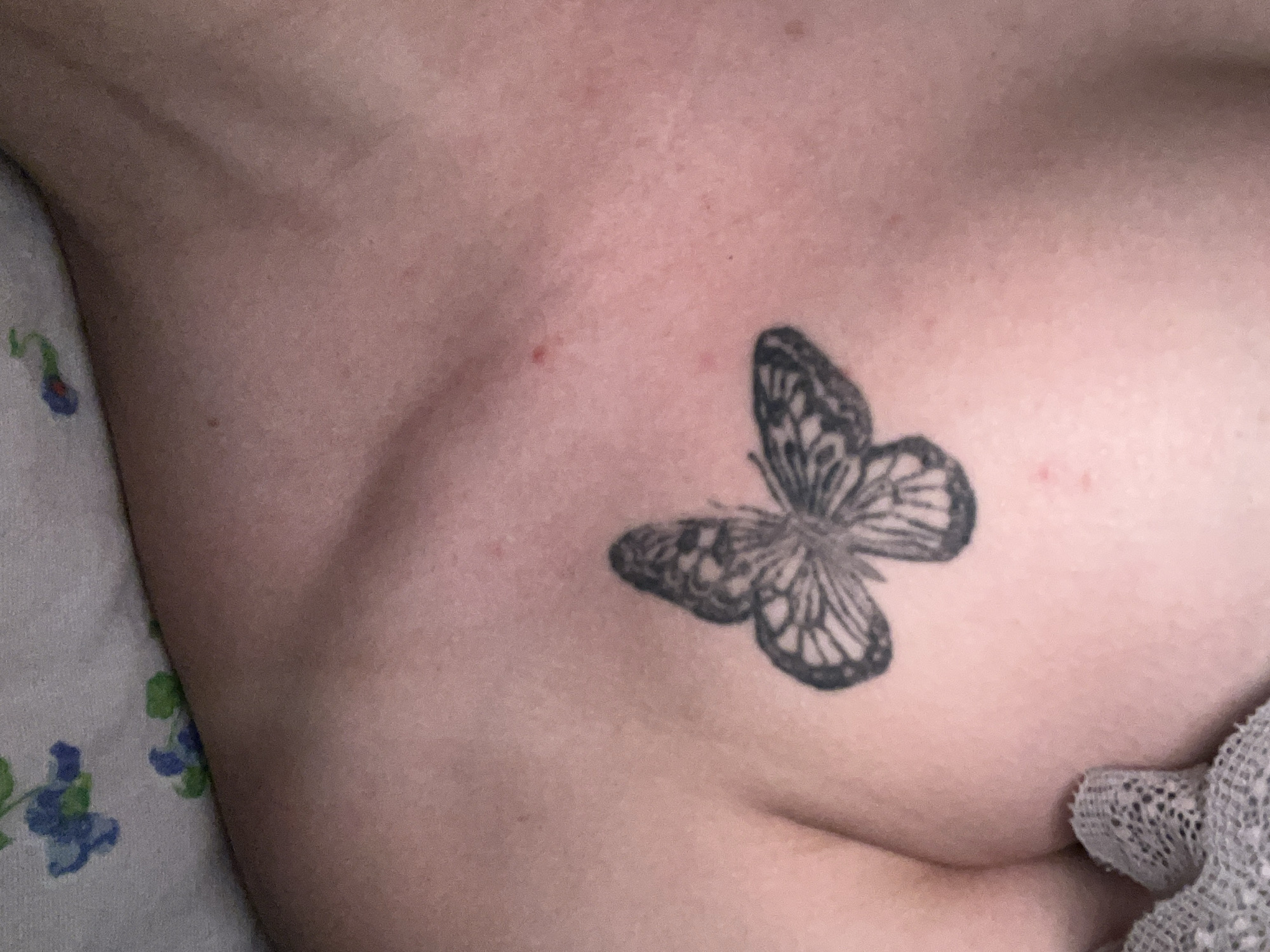 The image shows a black and grey tattoo of a butterfly on a person's skin, likely on the upper chest or shoulder area, given the curvature of the skin. The skin appears to have some pinkish undertones and minor blemishes. A textured grey fabric is visible in the upper right corner of the image, partially obscuring the skin. The lower part of the image shows a white surface with a floral pattern in blue and green. There is no indication of the city of Gorodets, Russia, or any specific urban environment.