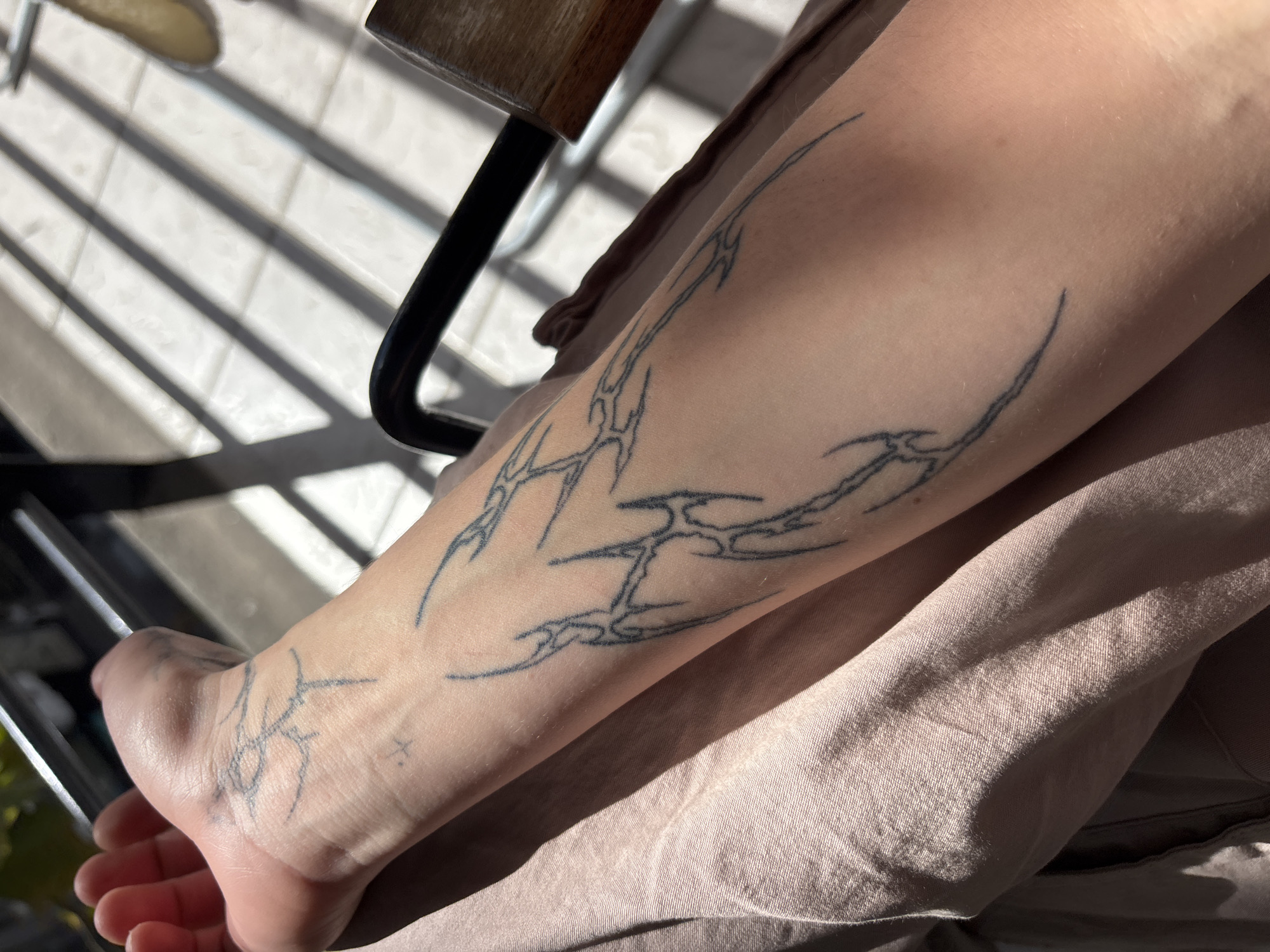 The image captures a close-up, downward-angled view of a person's lower leg and foot, illuminated by bright sunlight. The skin is a light to medium tone, adorned with multiple black ink tattoos in a tribal or abstract style. These tattoos feature sharp, intricate lines and shapes, extending from the top of the foot and ankle area, along the side of the calf. A small, simple "x" symbol tattoo is also visible on the top of the foot, near the ankle.
The person is wearing light brown or beige fabric clothing, possibly made of linen or cotton, which is visible in the lower right and background, showing soft folds and creases.
The scene is brightly lit, suggesting a sunny day, with strong, distinct parallel shadows cast across a light-colored tiled floor. These shadows, likely from railings or blinds, create a striped pattern on the ground and partially fall across the limb and clothing. A dark metal furniture leg, possibly from a chair, and a wooden surface are visible in the upper left corner, indicating an outdoor setting like a balcony or patio, or an indoor space with large windows. The overall impression is one of leisure in a sunlit environment. The location is Buenos Aires, Argentina.