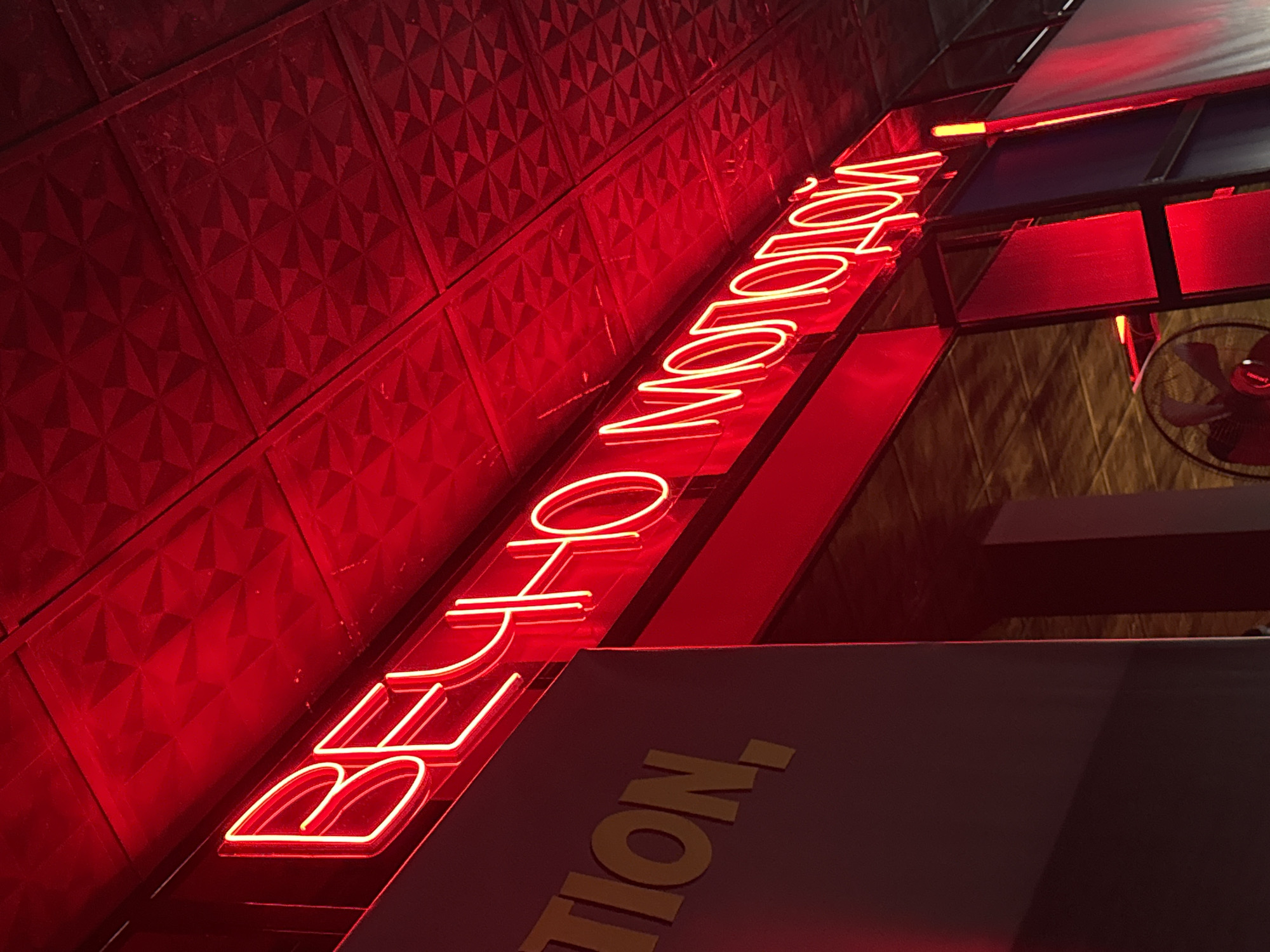 This is a low-angle, medium shot of a neon sign in a dimly lit setting. The sign is red and appears to be in Cyrillic script. The text reads "ВЕЧНО МОЛОДОЙ" which translates to "Eternally Young". The sign is mounted on a textured red wall and is illuminated by its own neon glow. There are also other elements visible in the frame, including a ceiling with what looks like fabric panels and metal framing, and a fan in the upper right corner. A dark surface with yellow text is visible in the lower right portion of the image, though the text is only partially legible, showing what appears to be "TION". The overall impression is of a retro or club-like atmosphere, possibly a bar or entertainment venue. The lighting suggests it is nighttime. The location context provided is Karon, Thailand.