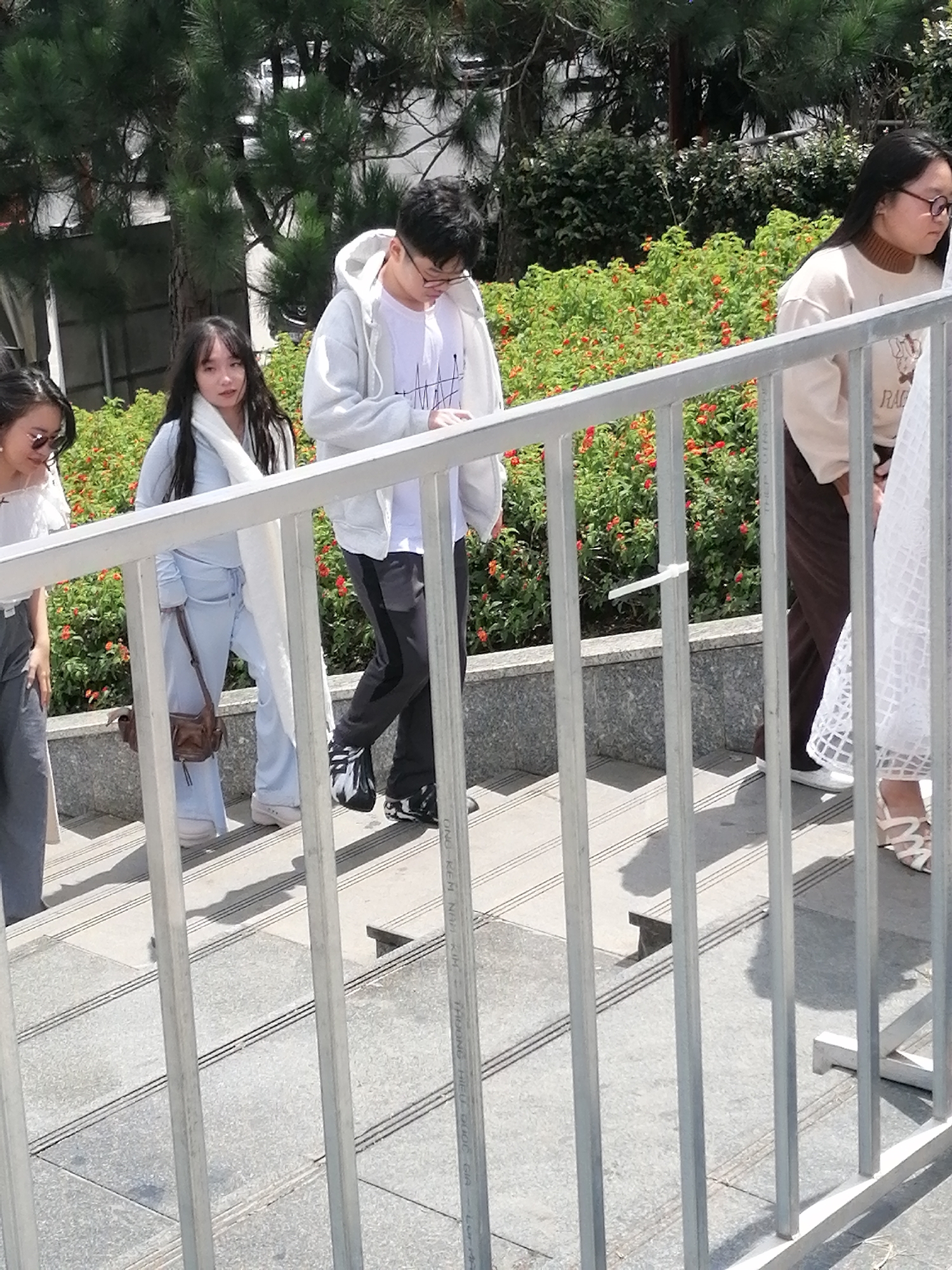 Four young adults, two male and two female, are walking up a set of outdoor stone stairs, partially obscured by a metal railing. The group is in a bright, outdoor environment with lush green foliage and small orange flowers visible behind them. The people are dressed casually, with one female wearing a light blue tracksuit and a white scarf, and another female in a beige sweater. The male on the right wears a light grey hooded sweatshirt and dark pants with a black stripe. The person on the far left is only partially visible, wearing sunglasses and a white top. The scene appears to be a public area, possibly near a park or garden, given the surrounding vegetation and stone construction. The lighting suggests it is daytime, with shadows cast on the stairs. The presence of a metal railing and the stone stairs indicates an urban or semi-urban setting. The image does not provide specific clues to pinpoint a location within Da Lat, Vietnam, but the general ambiance suggests a warm climate and a well-maintained public space.