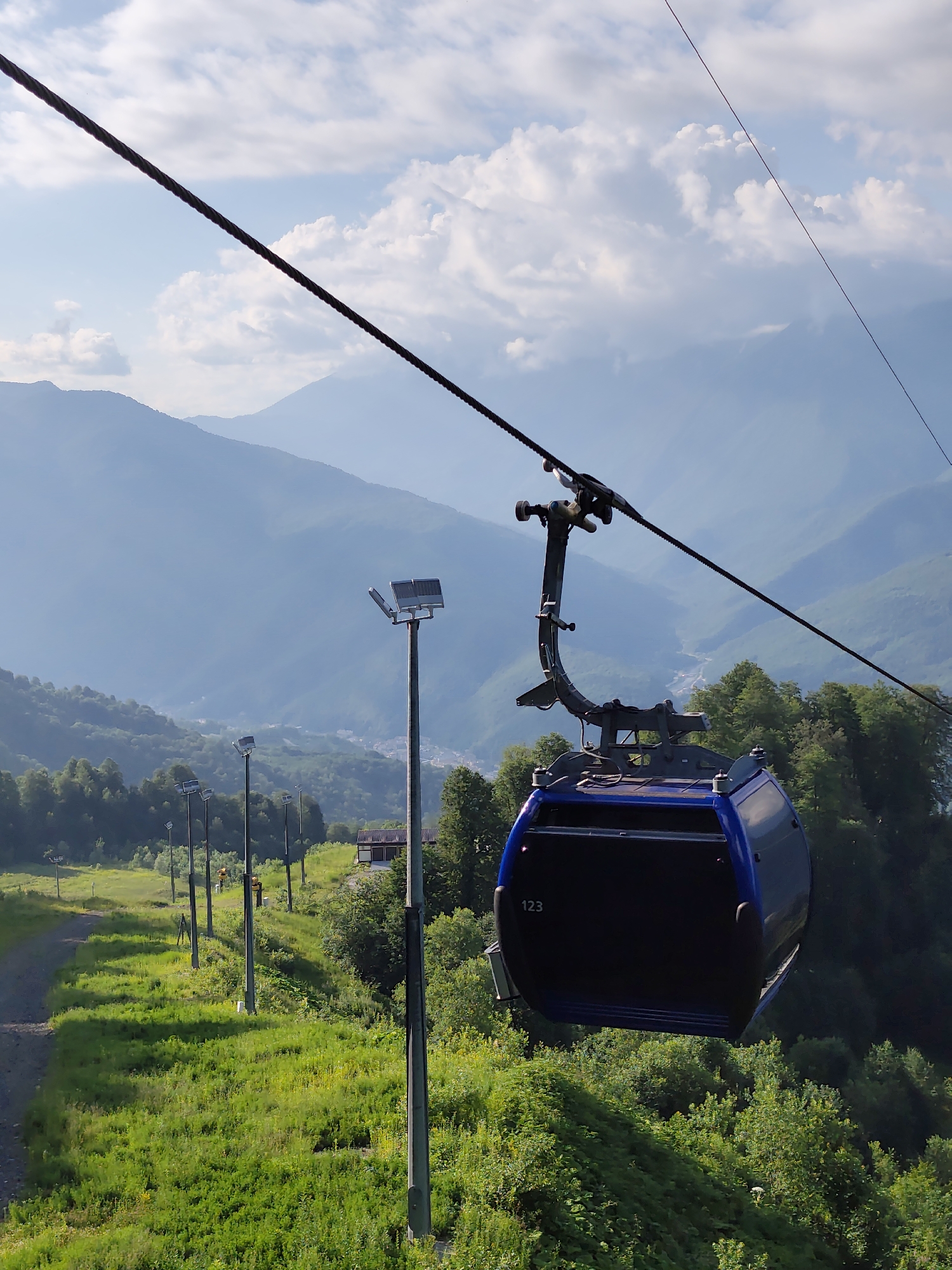 This is a vertical, eye-level shot of a blue cable car, with the number "123" visible on its side, suspended from a thick cable against a hazy blue sky filled with white clouds. The cable car is in the foreground, slightly to the right. Below and behind the cable car are rolling, tree-covered mountains and a green, grassy slope with several tall, grey poles topped with spotlights. A dirt path is visible on the left side of the slope. In the distance, layers of mountains fade into a soft, blue haze, and a small town or village is nestled in a valley. The overall impression is of a scenic mountain landscape, likely a resort area, in Estosadok, Russia.