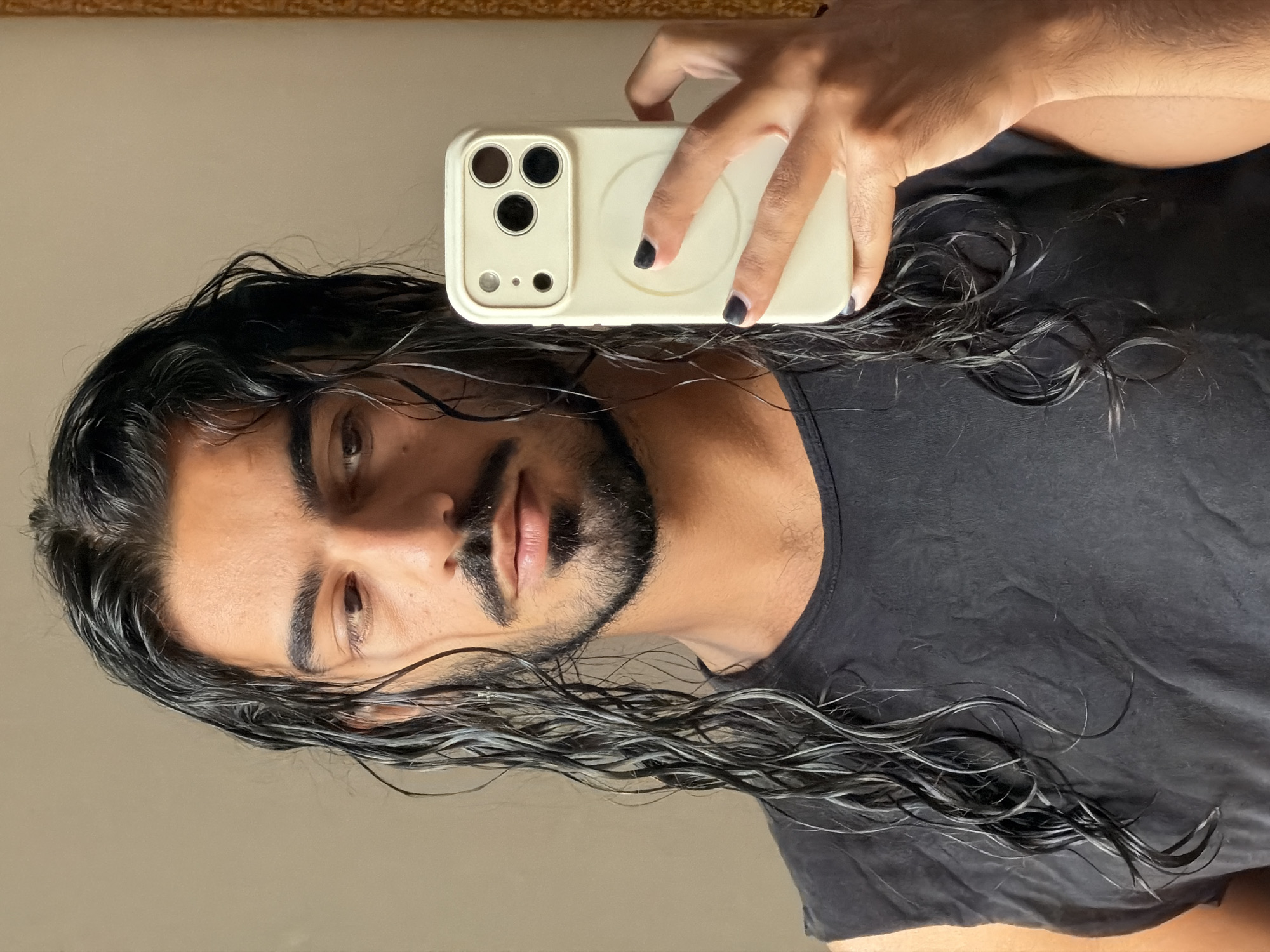 The media file shows a close-up, mirror selfie of a person with long, wet, dark hair and a beard. The person is wearing a black t-shirt. They are holding a light-colored phone with a camera array on the back. The background is a plain wall, reflecting the scene. The lighting suggests it is daytime. The image appears to be taken indoors. The person's hair is damp and styled in loose waves. The expression on their face is neutral. The overall mood is casual and informal. No visible text is present. The location context is North Kuta, Indonesia.