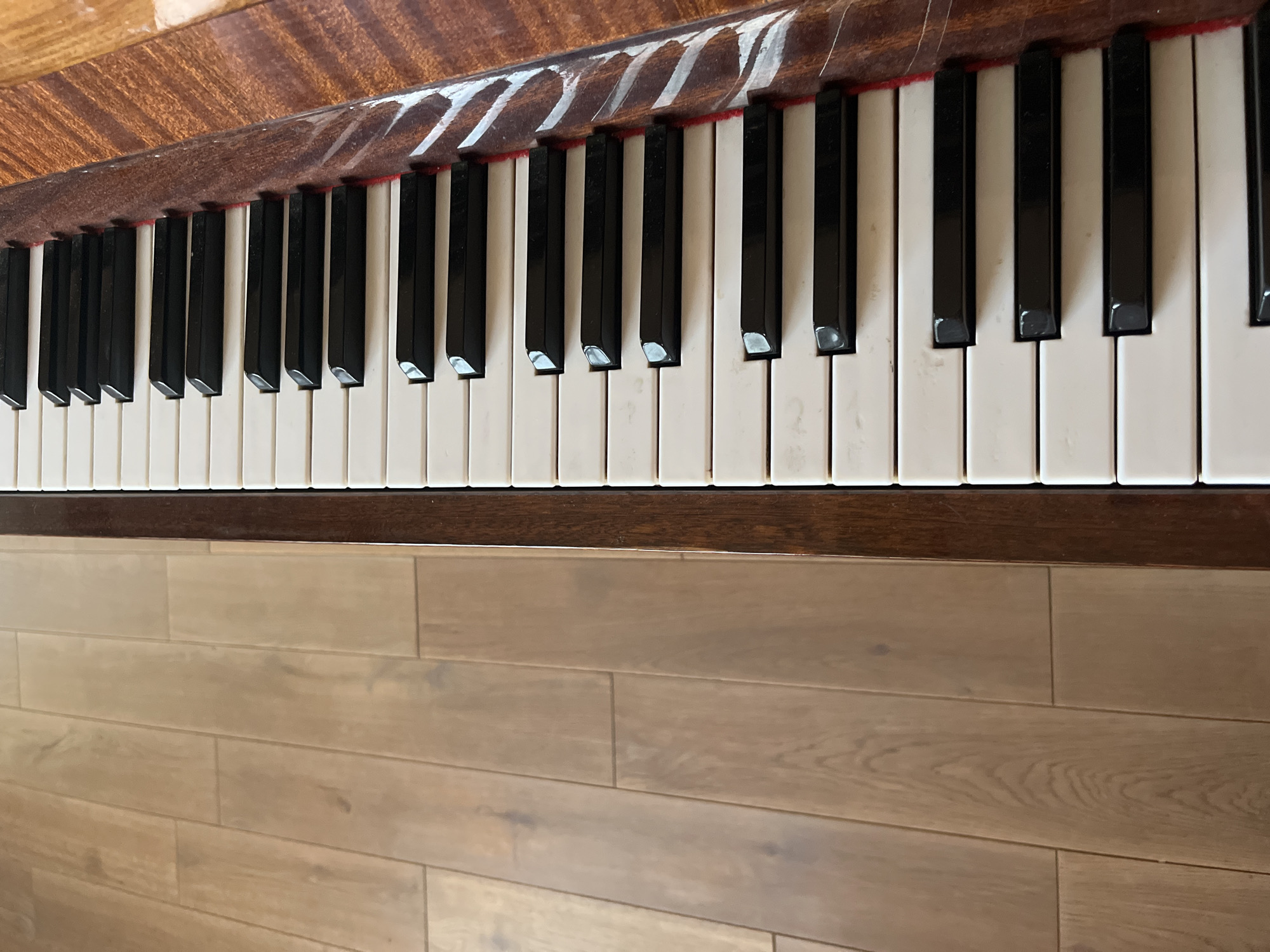 A close-up, vertical, eye-level shot captures a portion of a piano keyboard positioned on a wooden floor. The piano keys, white and black, are arranged in their typical alternating pattern, with the black keys slightly shorter and narrower than the white keys. To the left of the keyboard, the wooden floor is visible, composed of light brown planks laid horizontally. The texture of the wood grain is discernible. The right edge of the image shows the polished wooden casing of the piano, featuring a reddish-brown hue and a patterned grain. The scene appears to be indoors, with natural lighting suggested by the soft illumination. No people or actions are depicted. The location is not specified in the image itself.