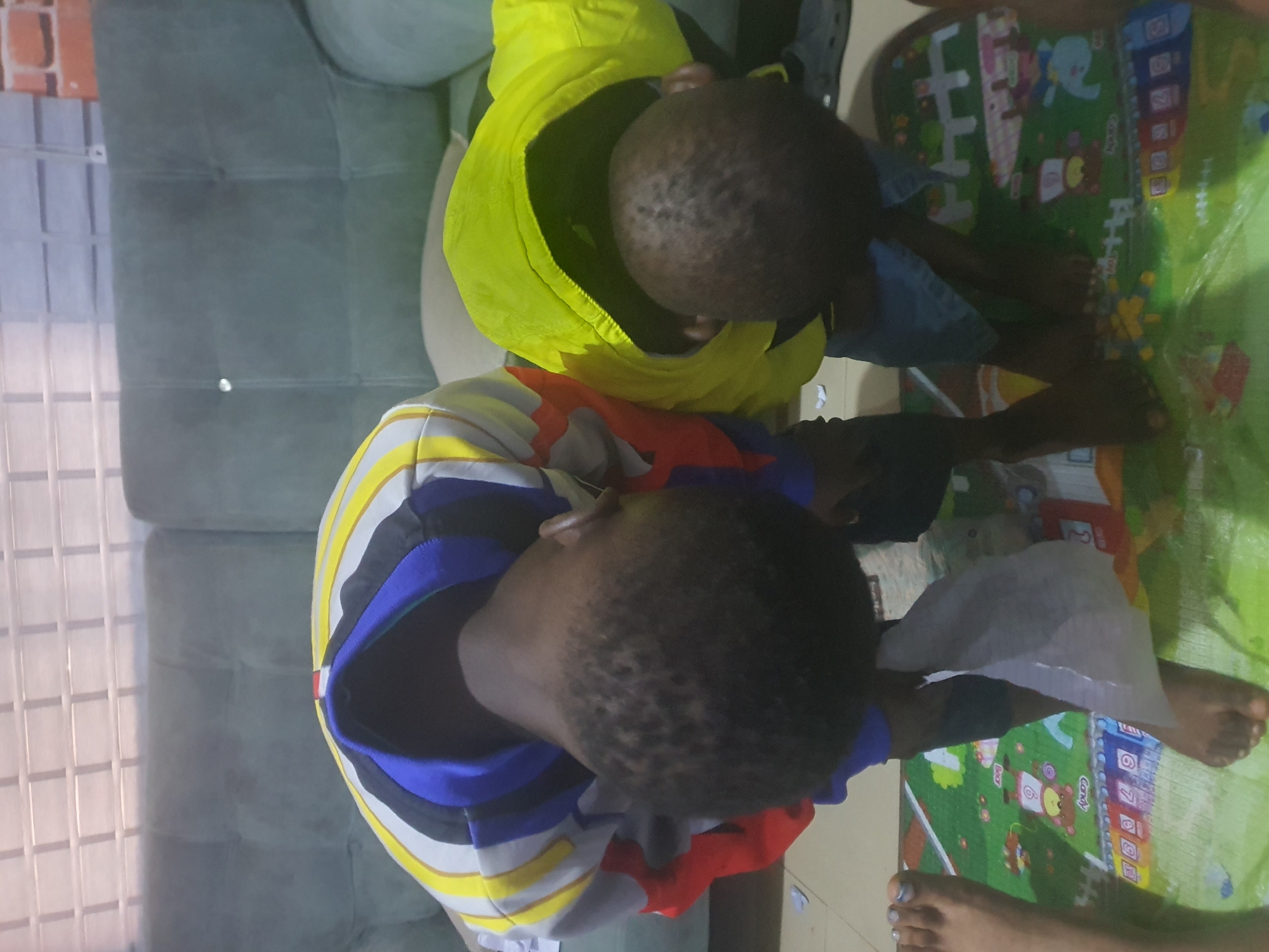 Two children are bent over, facing away from the camera, on a colorful playmat. The child on the left wears a jacket with horizontal stripes of yellow, white, blue, and red. The child on the right wears a bright yellow hooded jacket. Both children have short, dark hair and appear to be engaged in an activity on the playmat. A person's feet are visible on the left side of the frame, with one foot wearing nail polish. The background shows a couch and a wall with horizontal paneling, suggesting an indoor setting, possibly a living room or play area. This scene is located in Jos, Nigeria.