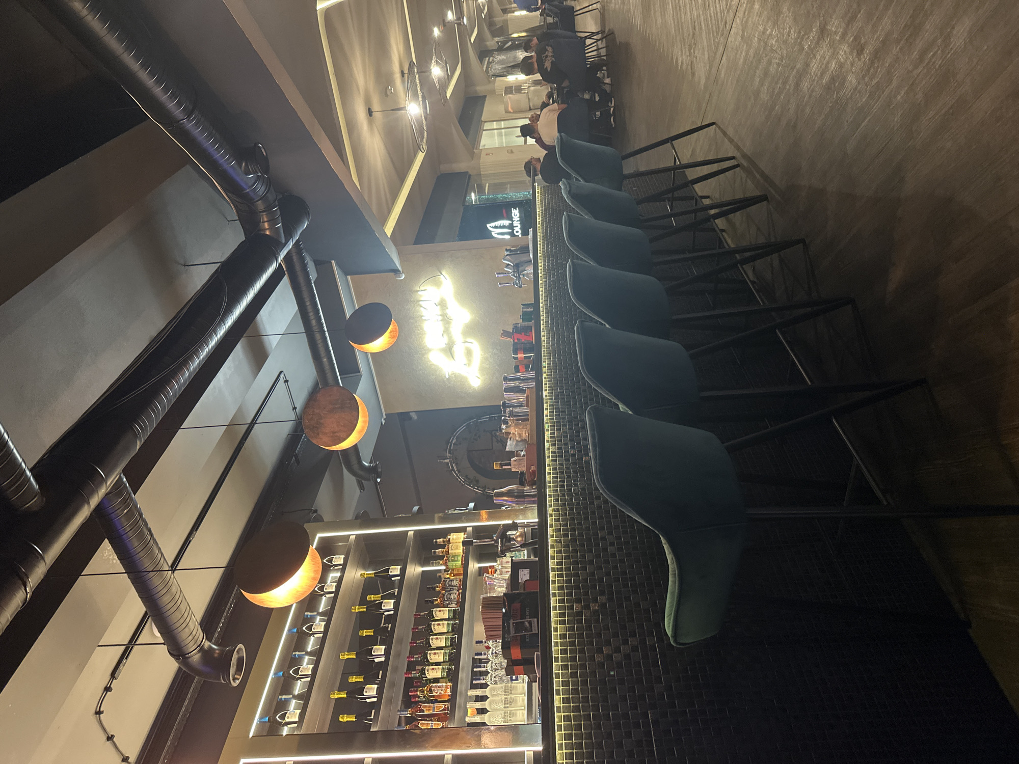 The image captures the interior of a bar, likely in Wrocław, Poland. The scene is dimly lit, and the focus is on a row of high bar stools facing a well-stocked bar counter. Behind the bar, shelves display an array of liquor bottles, illuminated by warm lighting. A neon sign, presumably the bar's name, hangs above the counter. The ceiling features exposed industrial-style ductwork and pendant lamps with a circular design, casting an orange glow. Further back, indistinct figures of patrons are visible.