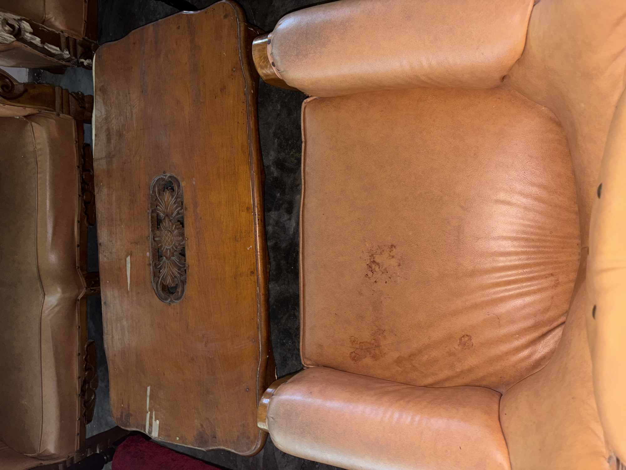 The image shows a close-up of a tan leather armchair and a wooden coffee table in a room. The armchair is worn and has several dark stains on the seat. The coffee table is also wooden and appears to be antique, with carved detailing in the center. The background is dark and out of focus. The image was taken in Beunyot, Indonesia.