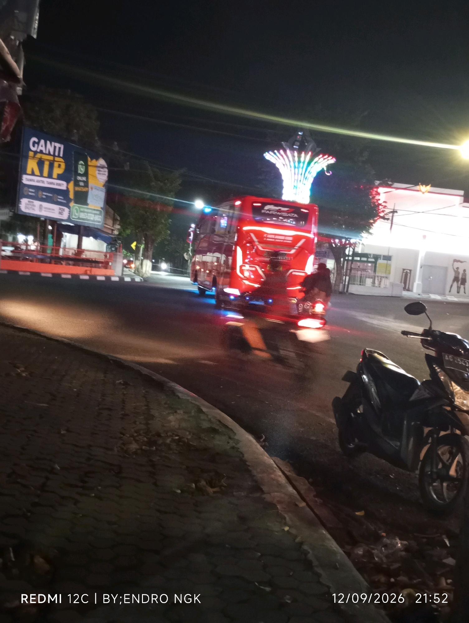 This is a nighttime street scene in Nganjuk, Indonesia. A red tour bus with illuminated signage is driving on the road. A motorcycle is parked on the right side of the image. On the left, there is a billboard advertising "GANTI KTP" (change ID card) and a business with outdoor seating. The road is paved, and there is a sidewalk with a tiled pattern. Trees are visible in the background. A decorative, brightly lit structure resembling a flower or fountain is also present. The bottom of the image contains a watermark with the text "REDMI 12C | BY:ENDRO NGK" and the date "12/09/2026 21:52".