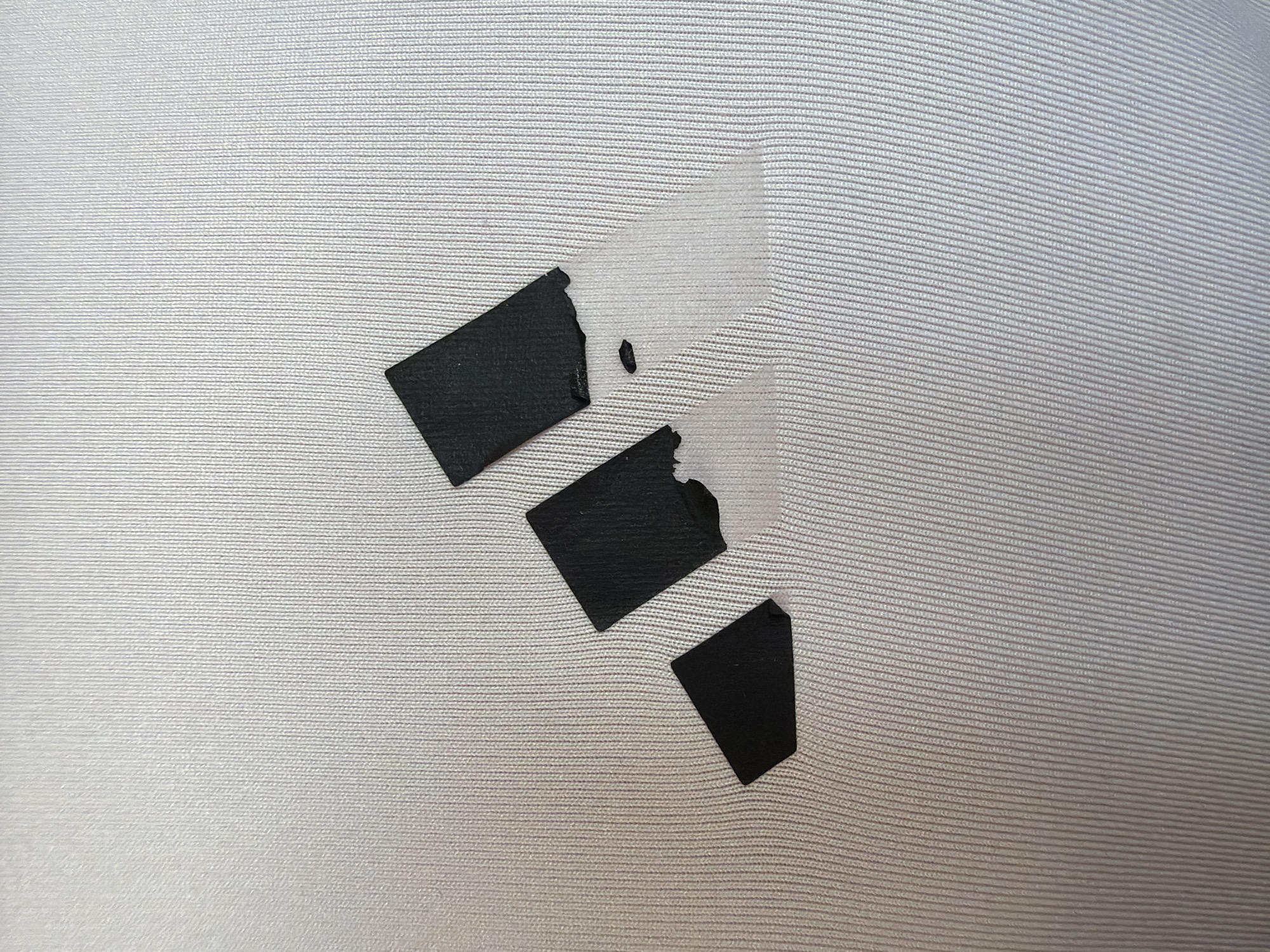 An Adidas logo on a white garment has peeled and is partially detached. The logo, composed of three black bars forming a triangular shape, is significantly damaged on its right side. The bars are peeling away from the fabric, with some areas of clear adhesive tape visible where the logo is detaching. The garment itself has a ribbed texture, with fine vertical lines. The image is a close-up shot, focusing entirely on the damaged logo and the fabric it is attached to. There are no people or background elements visible.