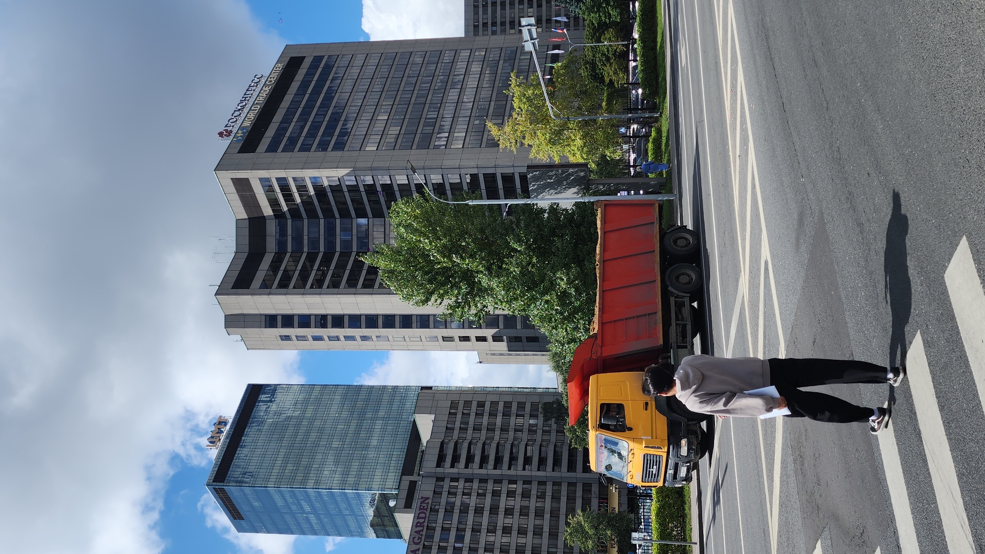 In Moscow, Russia, a person wearing a gray hoodie and black pants walks across a pedestrian crosswalk. The person is holding a white document. Behind the individual, a large yellow dump truck with a red bed filled with dirt is stopped on the road. In the background, towering buildings dominate the skyline. On the left, a modern glass skyscraper with "MGMMT" signage is visible. To the right, a taller, gray building with the "РОСКОНГРЕСС WORLD TRADE CENTER" logo stands prominently. Green trees and a street lamp are also present in the urban landscape under a partly cloudy sky.