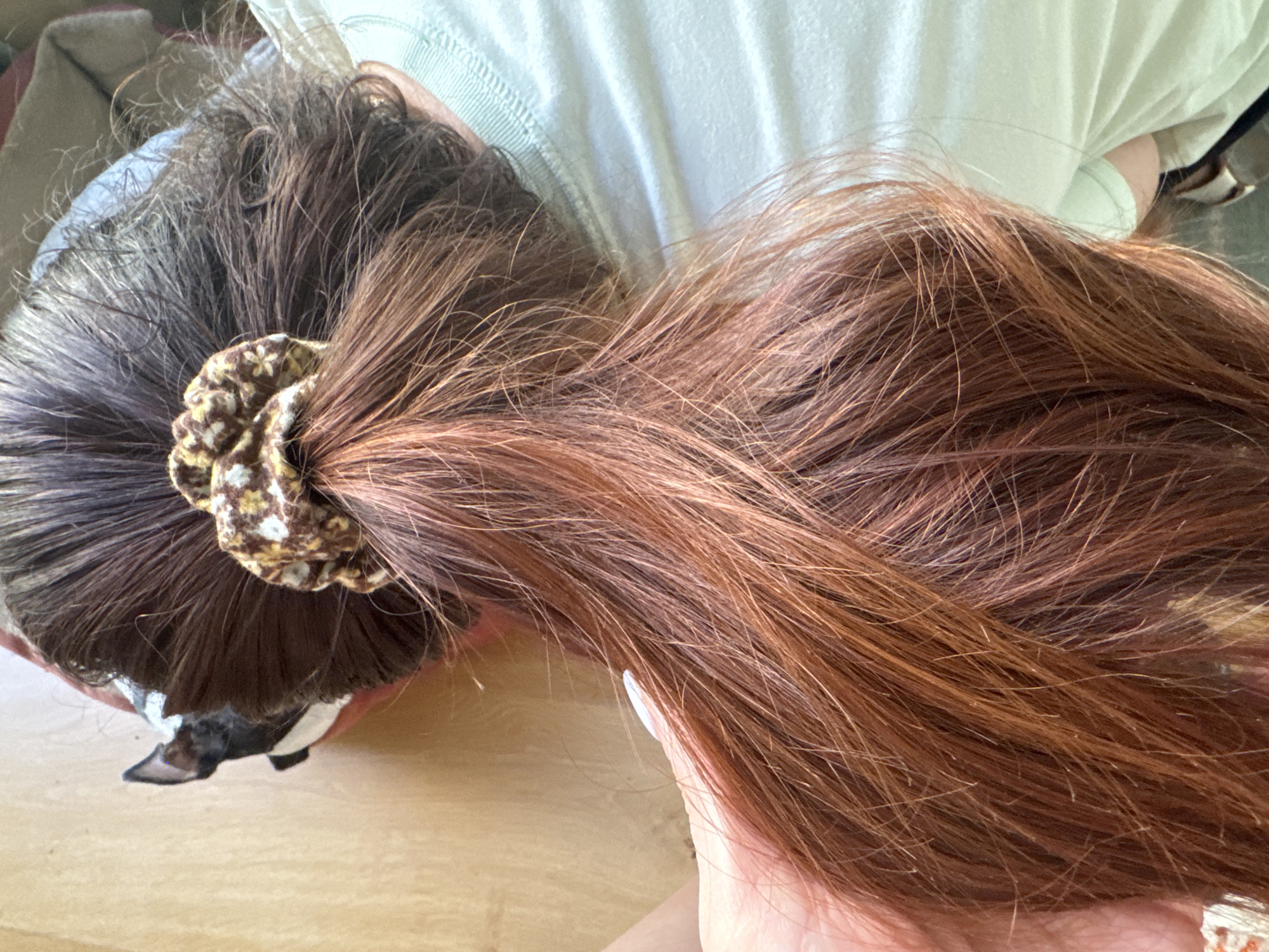 The image shows the back of a person's head, with their dark brown hair pulled back into a ponytail with a patterned scrunchie. The hair transitions to a reddish-brown color towards the ends. A hand with a light-colored fingernail is visible on the left side, touching the hair. The background is a light-colored wooden surface. There is no indication of the city of Murcia, Spain, in the image.