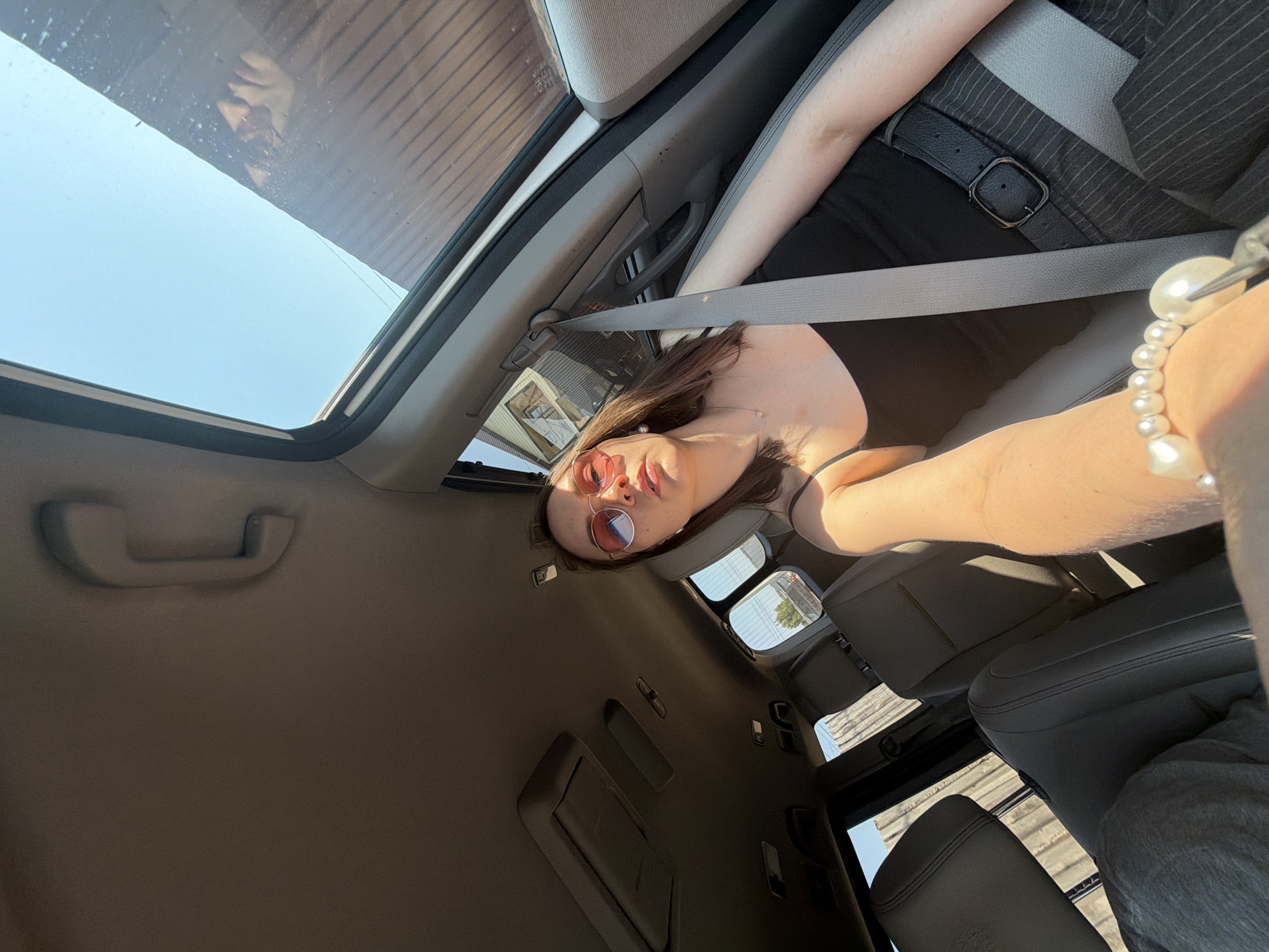 The image shows a person sitting inside a car, with a sunroof open. The person is wearing pink sunglasses and a black top. The car's interior is gray, and the person is wearing a seatbelt. There are buildings visible through the sunroof, suggesting an urban environment. The location is Chaikovsky, Russia.