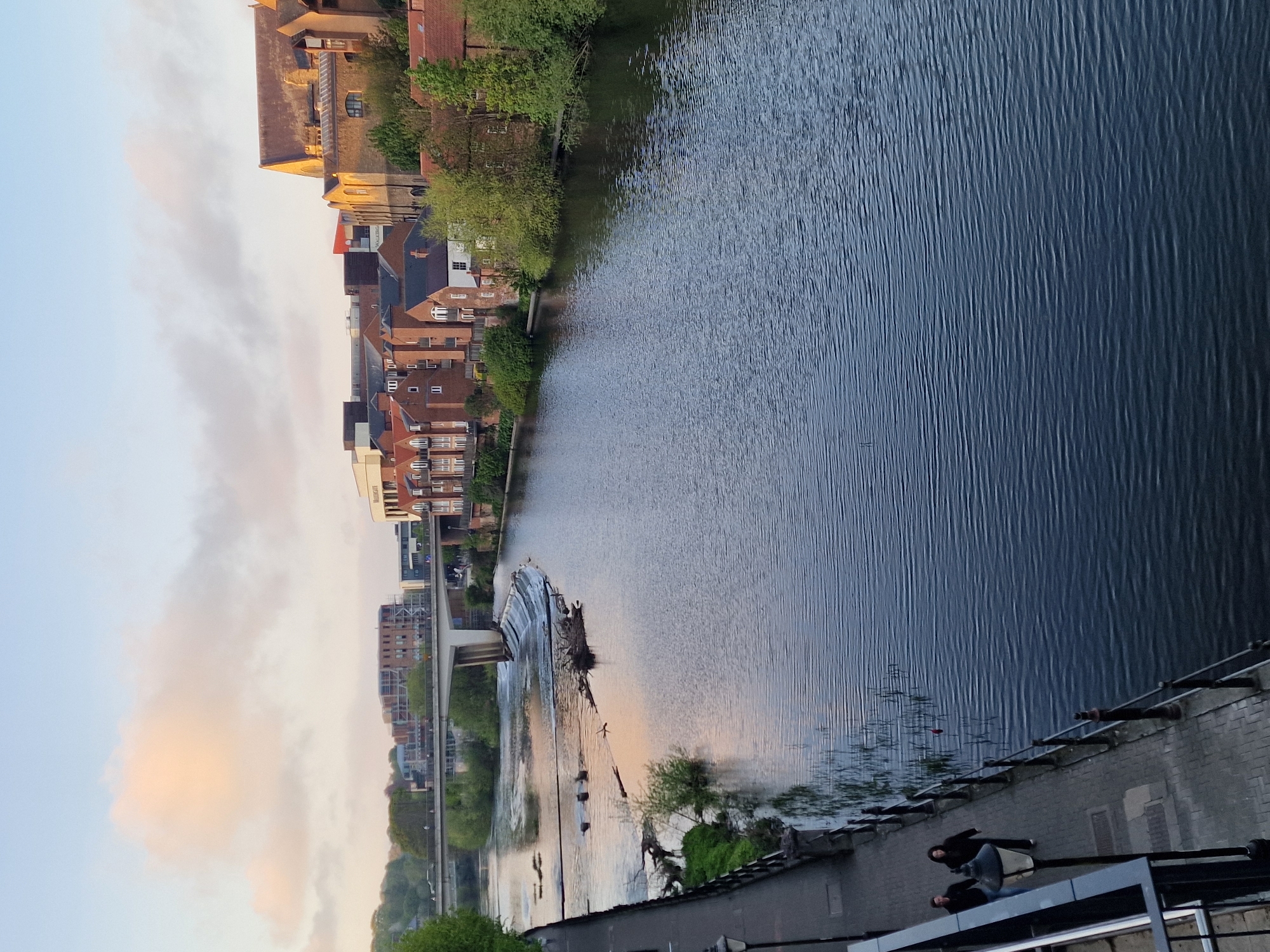 The image captures a serene waterside scene, likely of the River Wear in Durham, United Kingdom, seen during late afternoon or early evening. The overall composition is a wide landscape view with the river dominating the right side and buildings and sky on the left.
In the foreground, on the right side, two individuals are visible standing on a paved embankment or walkway alongside the river. One person is closer to the viewer, standing near a dark lamppost, while another is further back. The river itself, occupying the central and right portions of the image, is dark blue with numerous ripples across its surface, suggesting a gentle flow or slight breeze. Further into the river, a weir or small waterfall is visible, creating white foam and collecting several large logs and debris. A modern bridge with concrete supports spans the river further downstream from the weir.
The left bank of the river is lined with a variety of buildings. Many are multi-story structures made of brick with reddish-orange tiled roofs, typical of older British architecture. Lush green trees and vegetation grow abundantly along both riverbanks, softening the urban landscape. The sky above is a pale, diffused blue, with soft, light-colored clouds, some showing warm, peachy hues, indicative of the gentle light conditions of dusk or dawn. No discernible text is visible anywhere in the image. The scene conveys a peaceful and calm atmosphere.