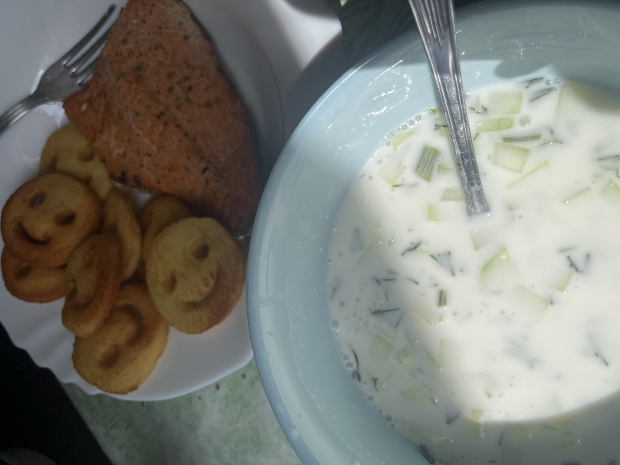 A top-down shot reveals a plate of food and a bowl of soup. The plate contains a piece of cooked salmon, a fork, and a small pile of smiley-face shaped potato patties. The bowl contains a creamy white soup with green herbs and diced cucumbers, and a metal spoon rests inside. The location and city are unknown.