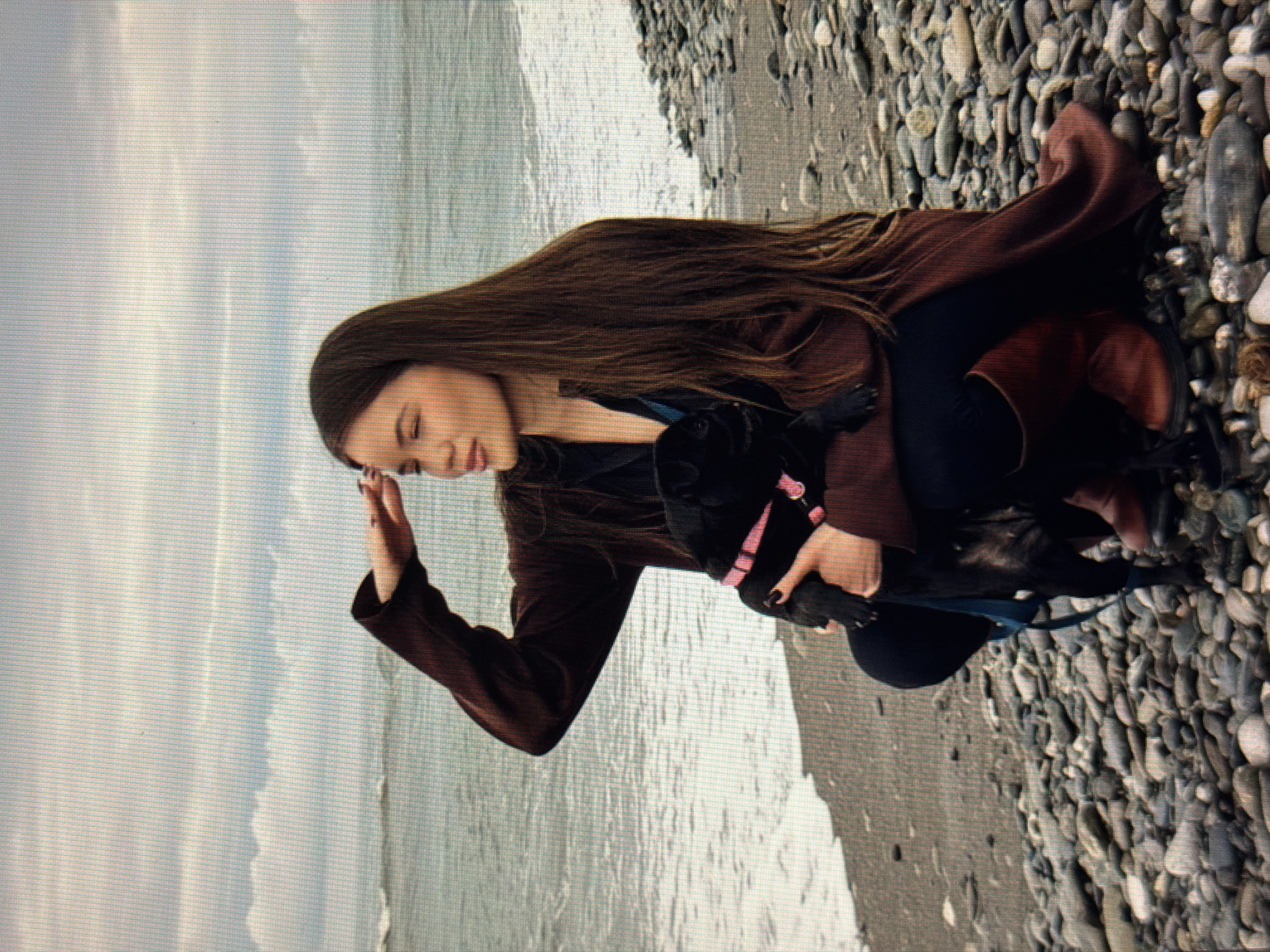 A young woman with long brown hair is crouching on a rocky beach, holding a black pug. She is wearing a dark brown coat and brown boots. The pug is wearing a pink collar. The woman is shielding her eyes with her hand, looking out towards the ocean. The sky is cloudy and the sea is choppy. The location is the shore of Sirius, Russia.