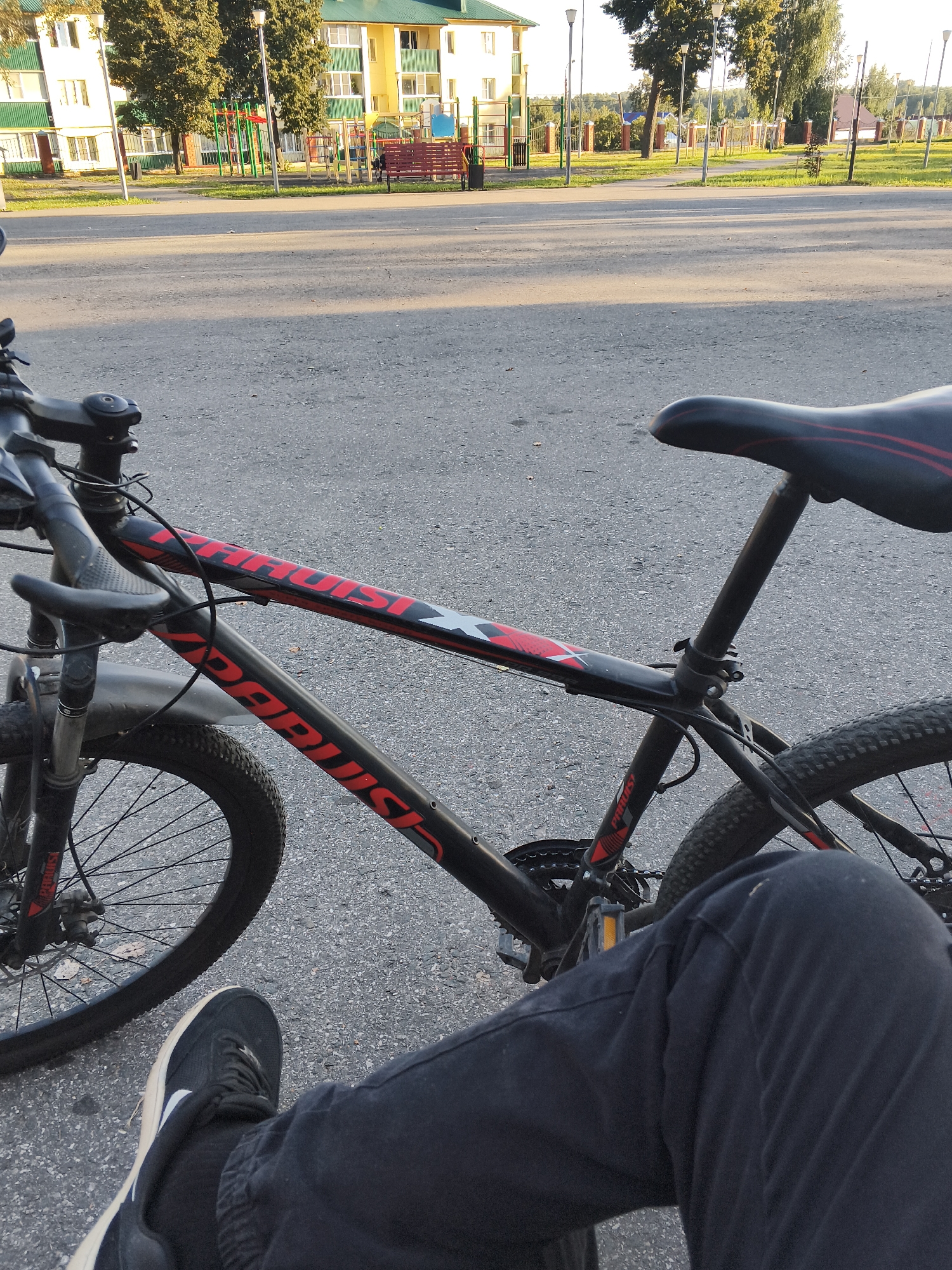 In Mokshan, Russia, a person's legs and feet, clad in dark trousers and black sneakers with white accents, are in the foreground, resting on an asphalt surface. The person is positioned next to a black mountain bike with red and white detailing on its frame. The bicycle's handlebars, seat, front and rear wheels, pedals, and part of the drivetrain are visible. The background reveals a city street with a playground featuring colorful equipment and benches, adjacent to apartment buildings with yellow and green facades. Tall trees line the street, and a fence separates the playground area from a grassy park. The scene is illuminated by natural daylight, casting subtle shadows on the asphalt.