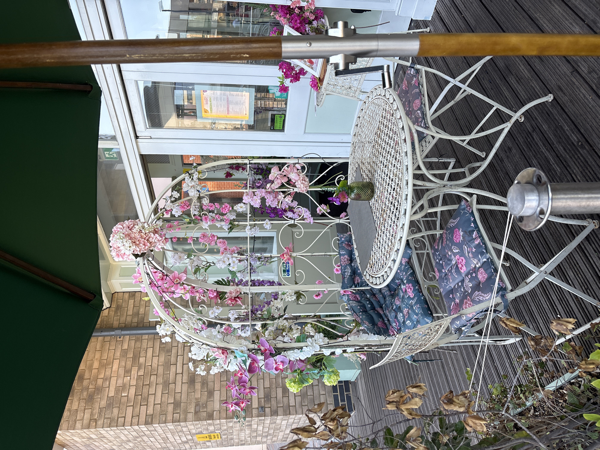 An outdoor seating area, likely a cafe or restaurant, is depicted in Chelmsford, United Kingdom. The scene is dominated by a white metal table and chairs arrangement, adorned with an abundance of artificial pink and white flowers cascading from a decorative white metal arch. The furniture is set on a dark wooden deck. A large green umbrella partially shades the area. In the background, a brick building with large glass doors and windows is visible, suggesting the establishment's entrance. A yellow warning sign is affixed to the brick wall. The overall impression is one of a charming, floral-themed outdoor space.