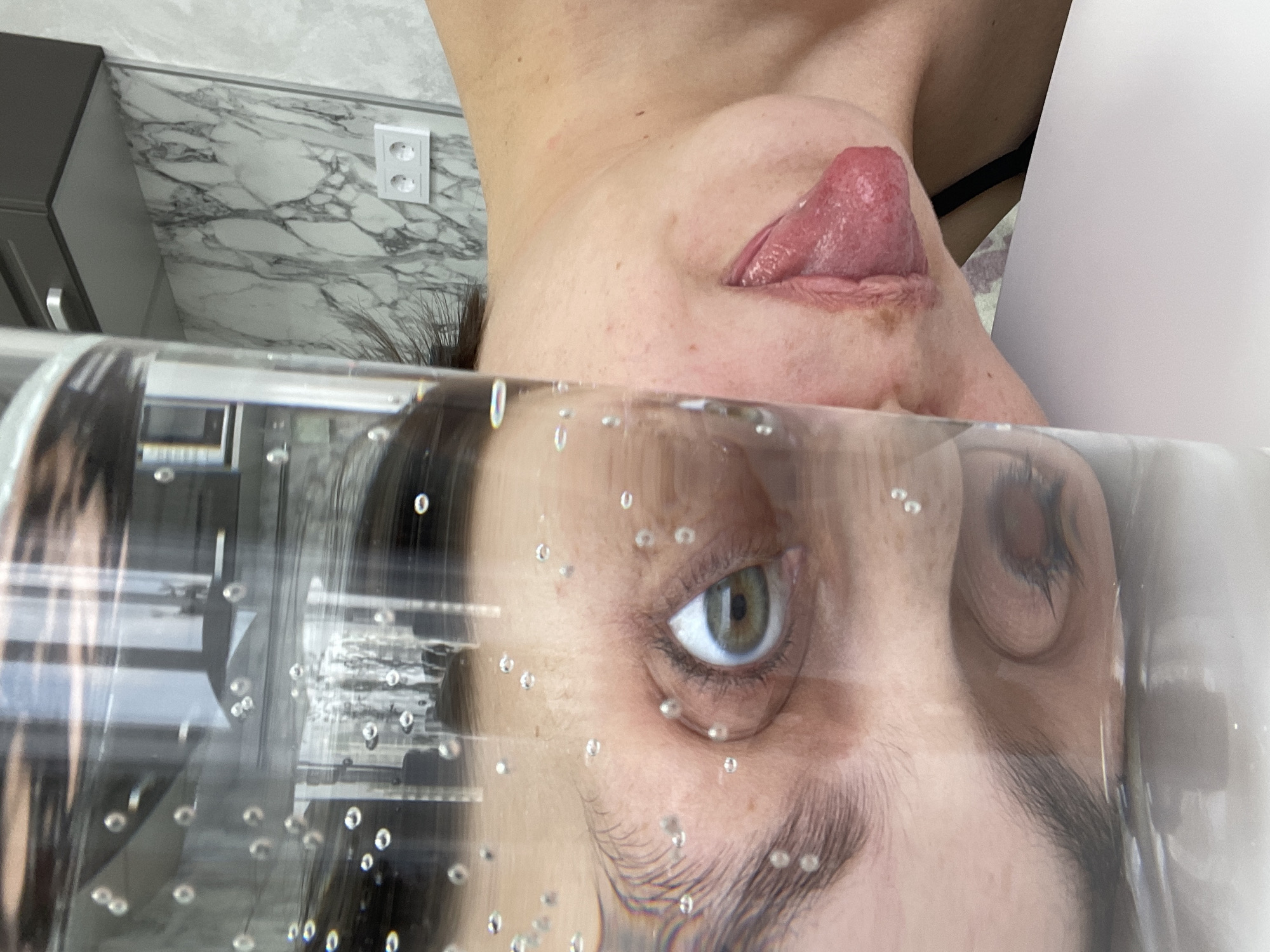 The image shows a person's face distorted through a clear container filled with liquid, possibly water. The person has one eye visible and open, with the other eye mostly obscured. Part of their tongue is protruding from their mouth. The background includes a marble-patterned wall, an electrical outlet, and a dark-colored cabinet. The photo is taken at an angle, appearing upside down. The city of Izhevsk, Russia, is not discernable from the information provided.