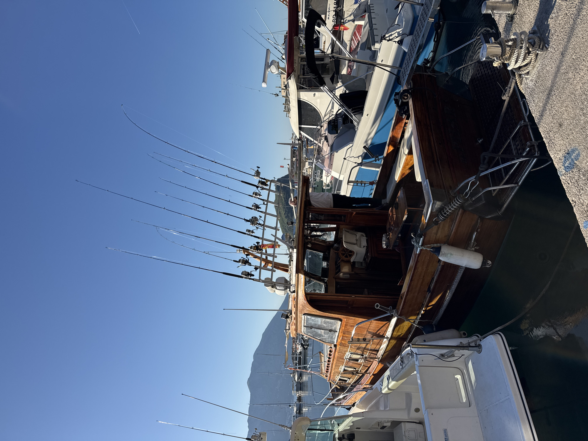 The image shows several boats docked side-by-side in what appears to be a marina. A wooden boat with numerous fishing rods extending upwards dominates the scene, moored next to a modern white boat. The clear blue sky and mountains in the background suggest a sunny day. Based on the mountainous backdrop, and presence of docked boats, the location is likely Budva, Montenegro.
