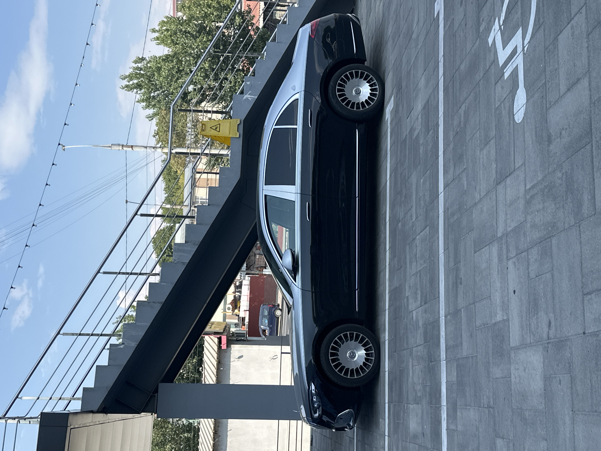 A black Mercedes-Maybach S-Class sedan with a silver roof is parked in a paved lot in Chișinău, Moldova. The car is positioned to the left of a grey outdoor staircase with metal railings. A yellow wet floor sign is placed at the bottom of the stairs. The ground is paved with large grey rectangular tiles, and white lines mark parking spaces, one of which is a disabled parking symbol. In the background, trees and buildings are visible under a clear blue sky. The scene appears to be outdoors during the daytime.
