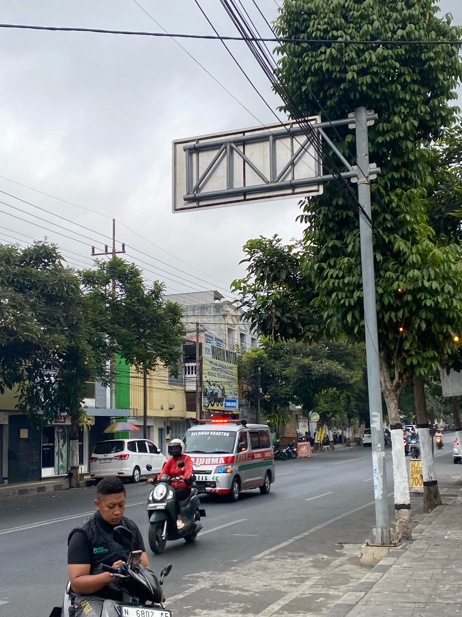A daytime, medium shot captures a street scene in an urban environment under an overcast sky. In the foreground, a man on a dark scooter is looking down at his phone, wearing a black vest over a t-shirt. He is positioned in front of a street sign. Behind him, an ambulance with flashing red lights is traveling down the street, followed by a woman on a dark green motorcycle wearing a red jacket and a white helmet. The street is lined with trees and buildings of varying architectural styles, some with storefronts. Power lines are visible crisscrossing the sky. The road has lane markings and a sidewalk. The overall impression is of a busy street with ongoing traffic.