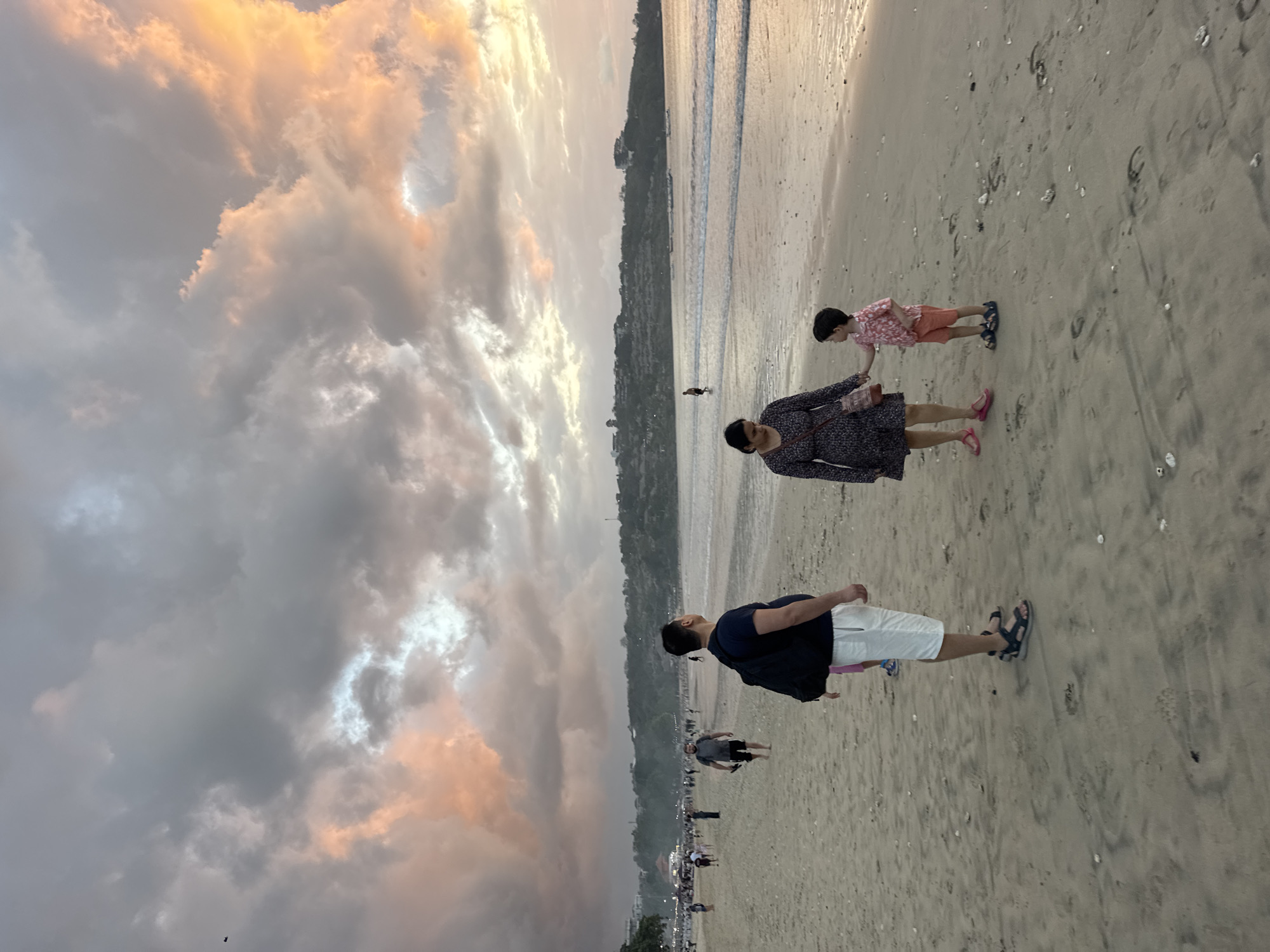 In Kuta Selatan, Indonesia, a family is captured on a sandy beach at dusk. The sky is filled with dramatic clouds illuminated by the setting sun, casting a warm orange and pink glow. In the foreground, a man stands with his back to the camera, wearing a dark t-shirt, white shorts, and sandals. He carries a black backpack. To his left, a woman in a dark floral dress holds the hand of a young boy wearing a coral-colored patterned shirt and shorts. They are facing the sea, with the ocean gently lapping at the shore. In the middle ground and background, numerous other people are visible walking along the beach, and a line of buildings and greenery stretches along the coastline. The beach is textured with sand and scattered seashells.