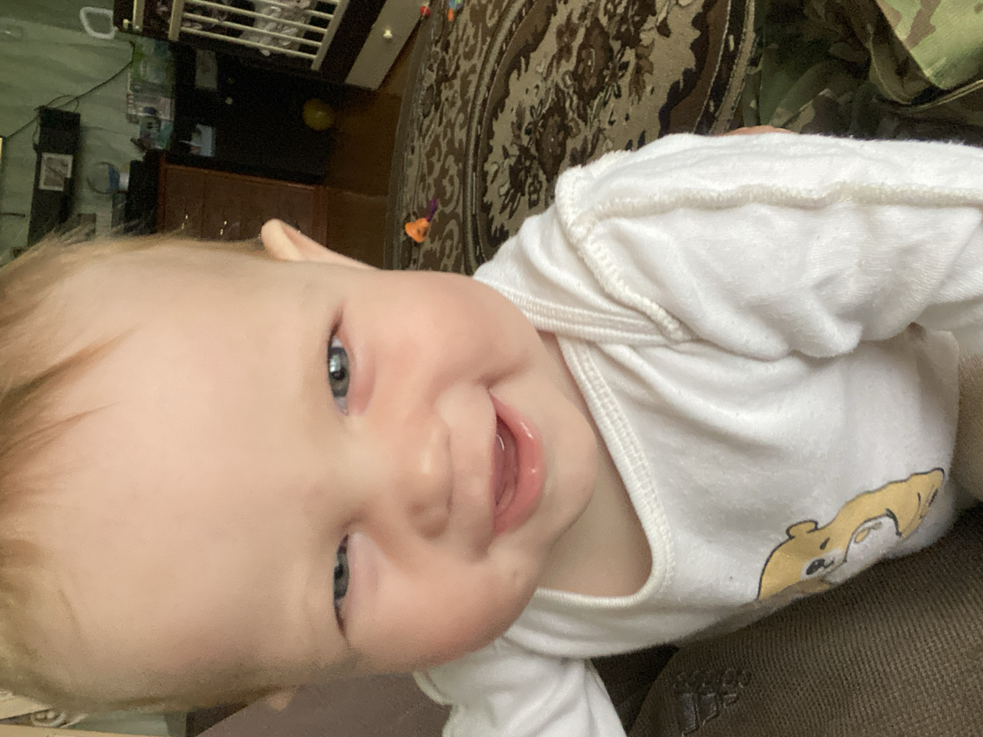 A close-up image depicts a fair-skinned infant with light hair and blue eyes, smiling broadly with a partially open mouth and visible tongue. The infant's right eye is open, while the left eye is partially closed, creating a winking or squinting expression. The infant wears a white long-sleeved onesie adorned with a partial yellow bear graphic on the left chest area. A brown fabric, possibly part of an adult's attire, is visible on the extreme left. The background indicates an indoor residential setting in Фоки, Russia. Visible elements include a dark wooden dresser or cabinet on the left, a white crib or playpen with vertical bars on the right, and a patterned rug covering the floor. An orange and purple toy, along with a yellow spherical object, are present on the rug. The wall behind the infant appears to be a muted green.