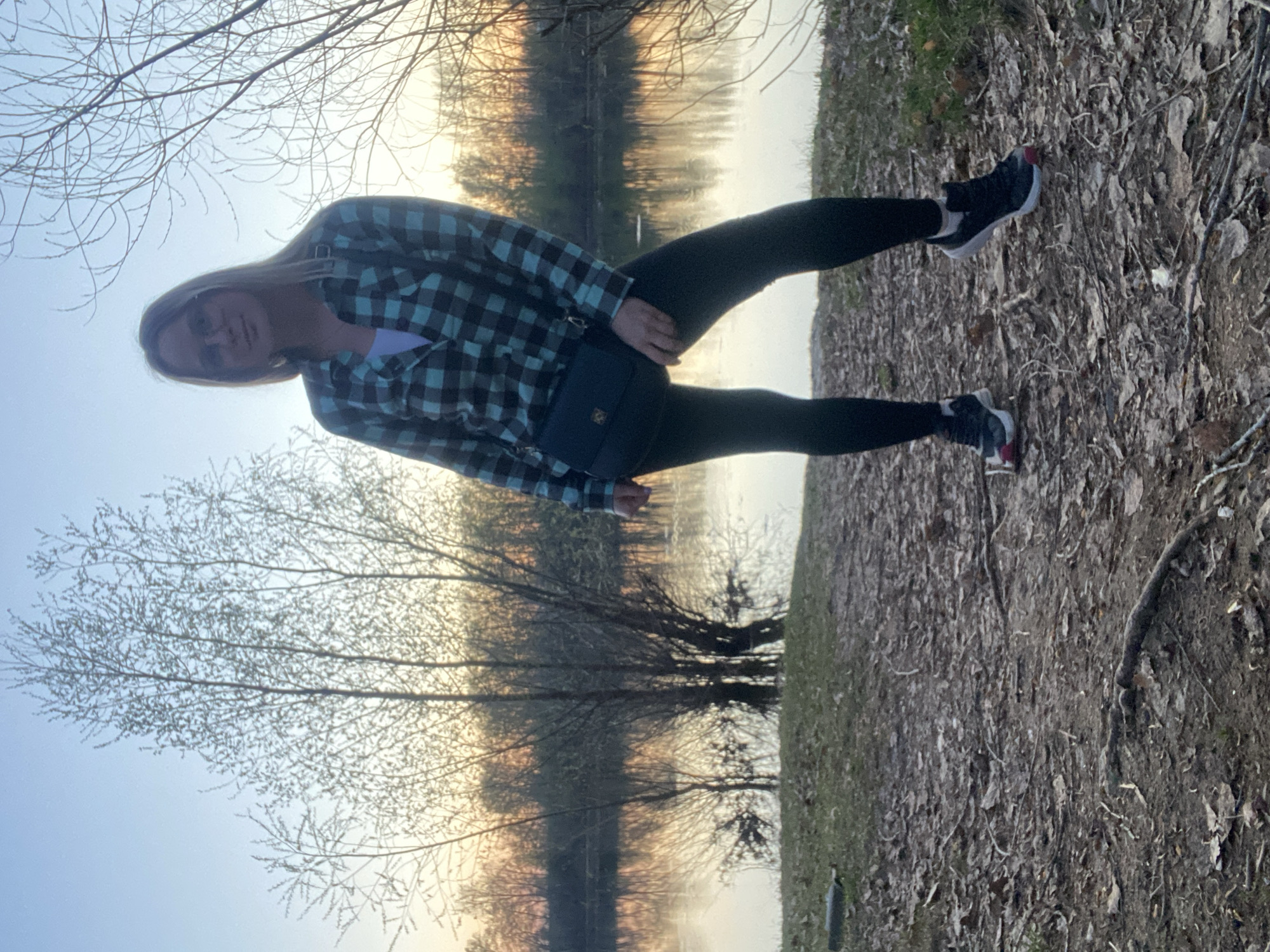 A woman stands on the edge of a path. She has long blonde hair and is wearing black leggings, black and white running shoes, and a plaid shirt. A black purse hangs at her hip. Behind her is a body of water that may be the Kama River. The picture might have been taken in Chaikovsky, Russia. There are bare trees along the bank of the river and the path the woman stands on. The ground is covered with dead leaves. The sky is light blue.