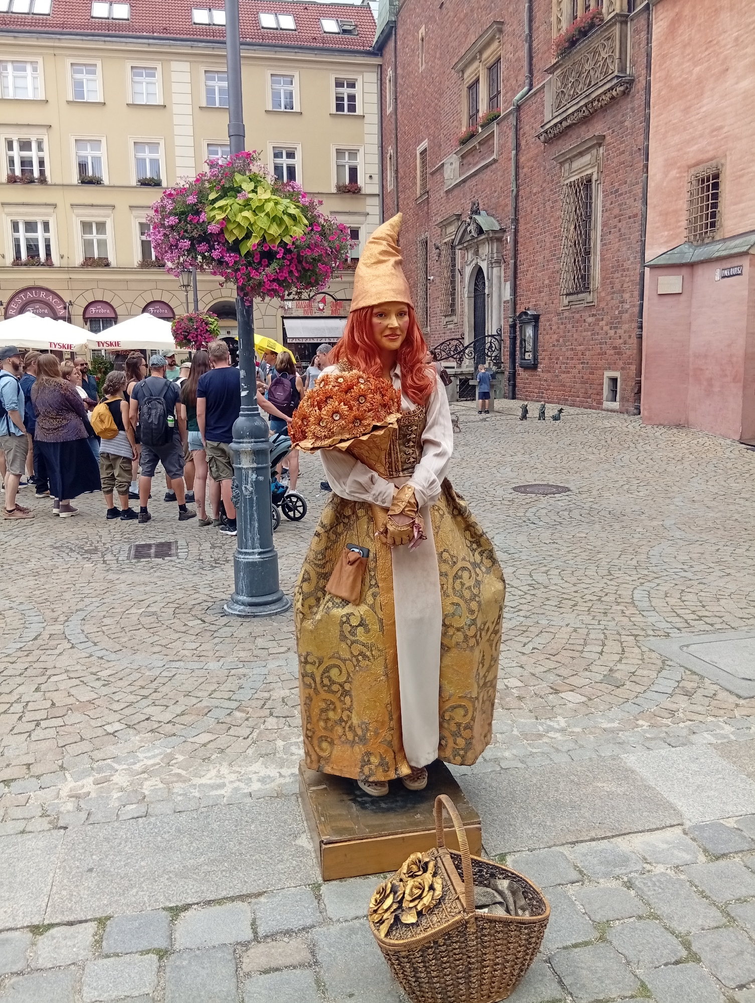 A street performer, painted gold with red hair and a tall conical hat, stands on a wooden platform on a cobblestone square in Wrocław, Poland. The performer wears a flowing gold and cream gown decorated with intricate patterns and holds a bouquet of orange flowers. A woven basket with fabric and decorative roses sits at the base of the platform. In the background, a crowd of onlookers gathers near white tents and a lamppost adorned with hanging flowers. Red brick buildings with arched windows and balconies line the square.