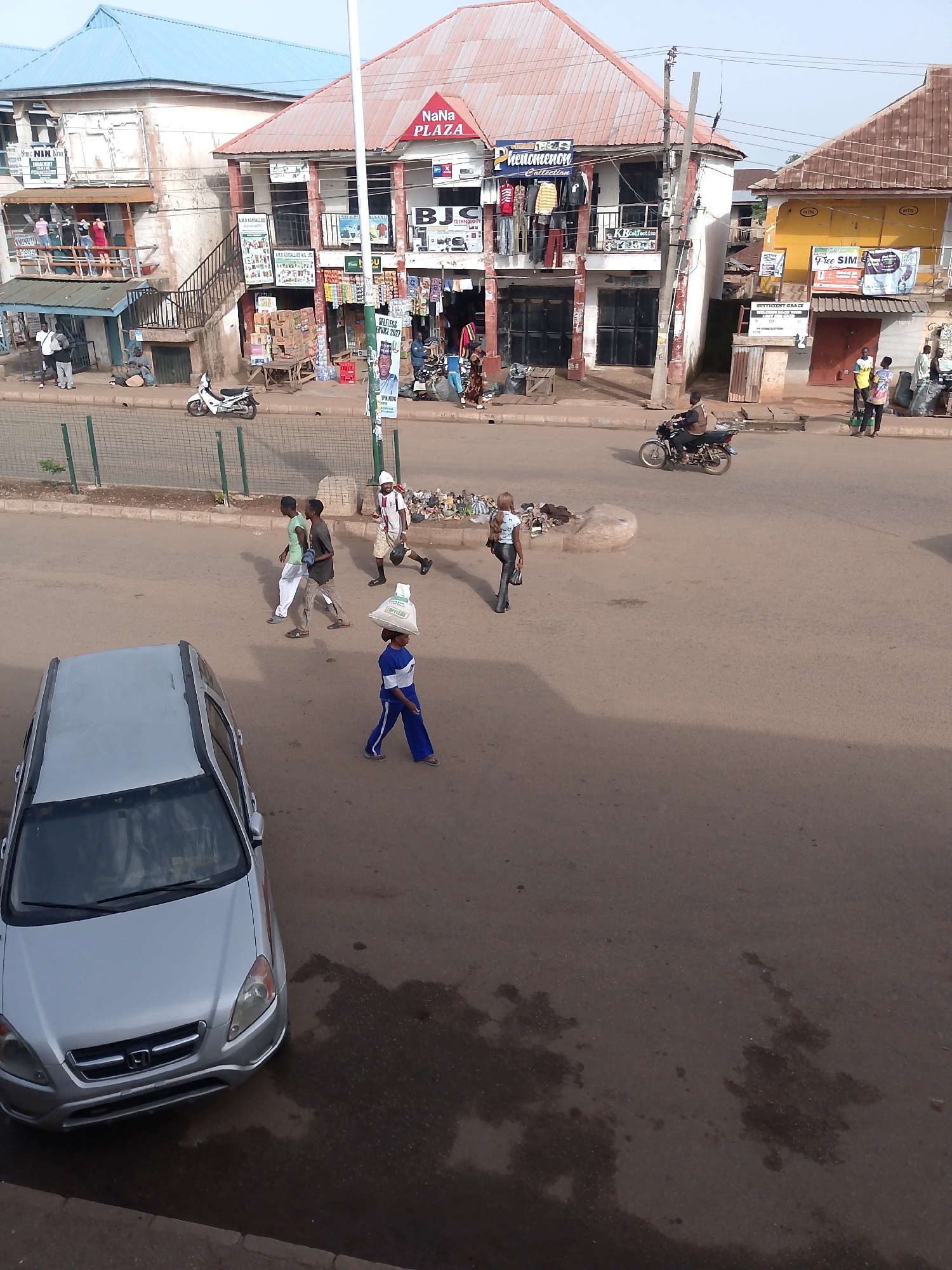The image depicts a street scene in Bukuru, Nigeria, with multiple buildings lining the road. In the foreground, a silver sedan is parked facing forward. To the right of the car, a young person wearing a blue and white outfit walks with a large bag balanced on their head. Further down the street, three young men walk together. On the right side of the road, a motorcycle with a rider is in motion. Various shops and businesses are visible in the background, including signs for "NaNa PLAZA" and "Phenomenon Collection". People are seen standing and walking around the shops. A white motorcycle is parked near one of the buildings. The environment appears to be an urban setting with paved roads and sidewalks.