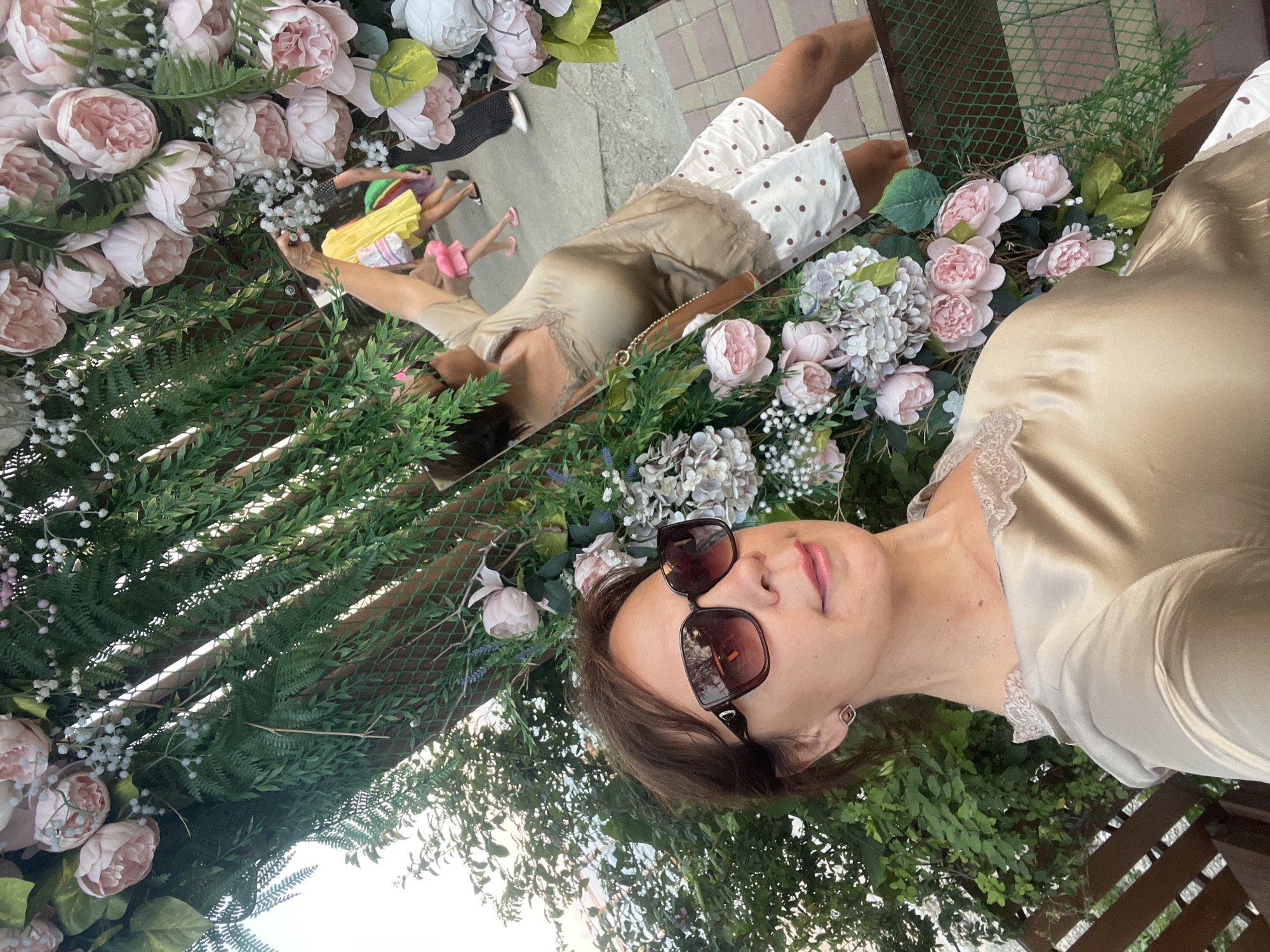 A woman with brown hair, wearing large sunglasses and a light-colored silk top with lace trim, is taking a selfie. She is positioned in the foreground, with her face and upper body visible. Behind her, a woman in a beige top and polka-dot shorts is reflected in what appears to be a mirror or reflective surface, her arm raised as if posing for a photo. In the background, other people are visible walking, including a person in a yellow dress and another in dark pants. The setting is outdoors, adorned with a lush arrangement of artificial flowers, including pink peonies and hydrangeas, and greenery, resembling a decorative archway or backdrop. The ground is paved with light-colored square tiles. The overall scene suggests a public event or a decorated area, possibly in Sochi, Russia, given the context.