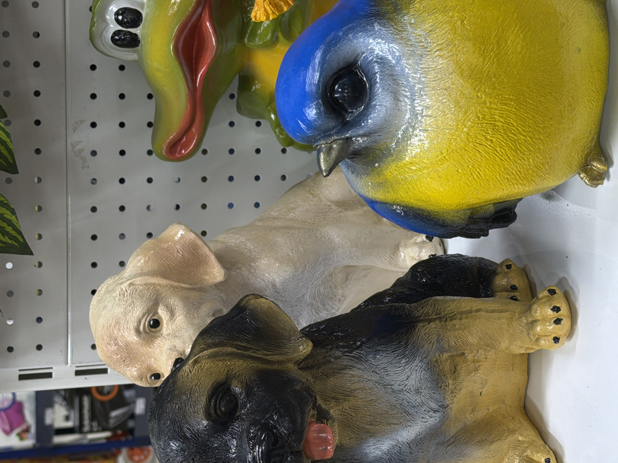 The image presents a close-up shot of a display of garden statues. From top to bottom, there is a green and red frog statue, a blue and yellow bird statue, a tan labrador statue, and a black and tan German shepherd statue. The statues are on a white wall with round holes, likely a display rack in a store. It is not possible to know the city of Perm, Russia from this image.