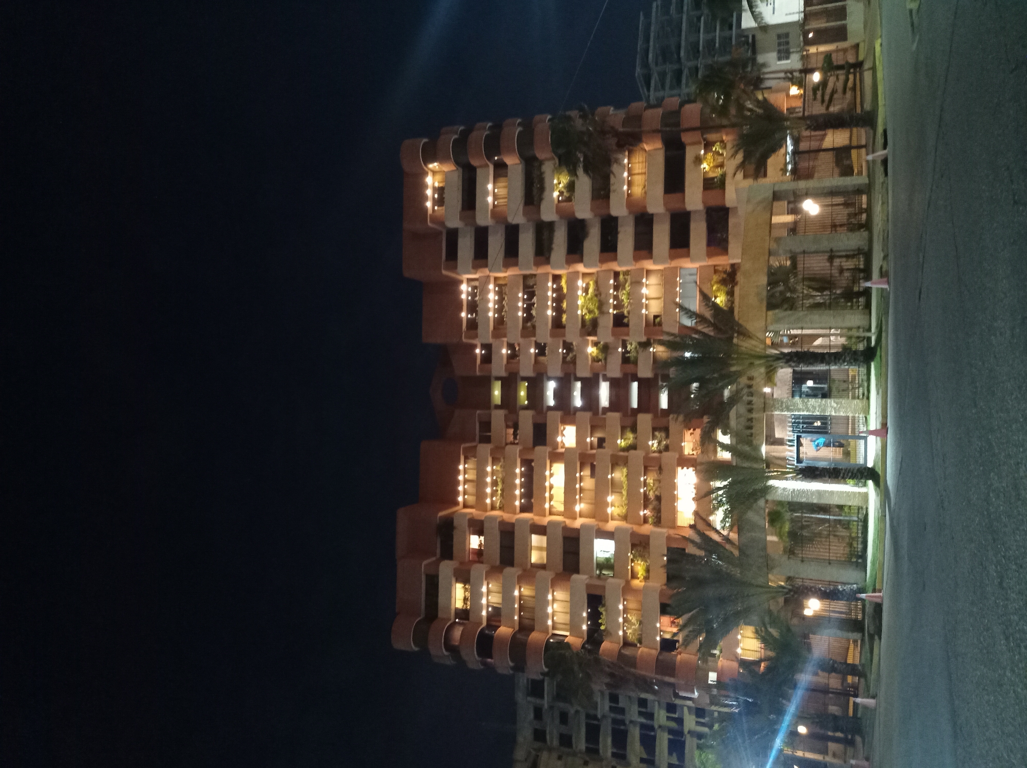 The image captures a multi-story building at night, viewed from an outdoor street or pathway. The sky is dark, indicating nighttime, and the scene is illuminated by artificial lights.
The main subject is a large, modern building, possibly a hotel or apartment complex, characterized by a distinct architectural style. Its facade features a repeating pattern of recessed balconies or window sections, many of which are lit from within, casting a warm, golden glow. Several balconies are also adorned with strings of small, bright lights, enhancing the festive or welcoming ambiance. Green foliage and plants are visible on some balconies, adding a touch of nature to the structure.
In front of the building, several tall palm trees are visible, their dark silhouettes contrasting with the illuminated facade. A paved street or pathway runs along the right side of the frame, lined with a dark metal fence or gate. Several orange traffic cones are intermittently placed on the pavement. Streetlights cast pools of light on the ground and parts of the building's lower levels.
On a stone pillar or wall near what appears to be the entrance, the word "ALEXANDRE" is partially visible, suggesting the name of the establishment. There are no people visible in the image. The overall impression is one of a well-lit, inviting establishment in a tropical or subtropical setting, consistent with its location in Porlamar, Venezuela. The weather appears clear, with no signs of rain or fog.
