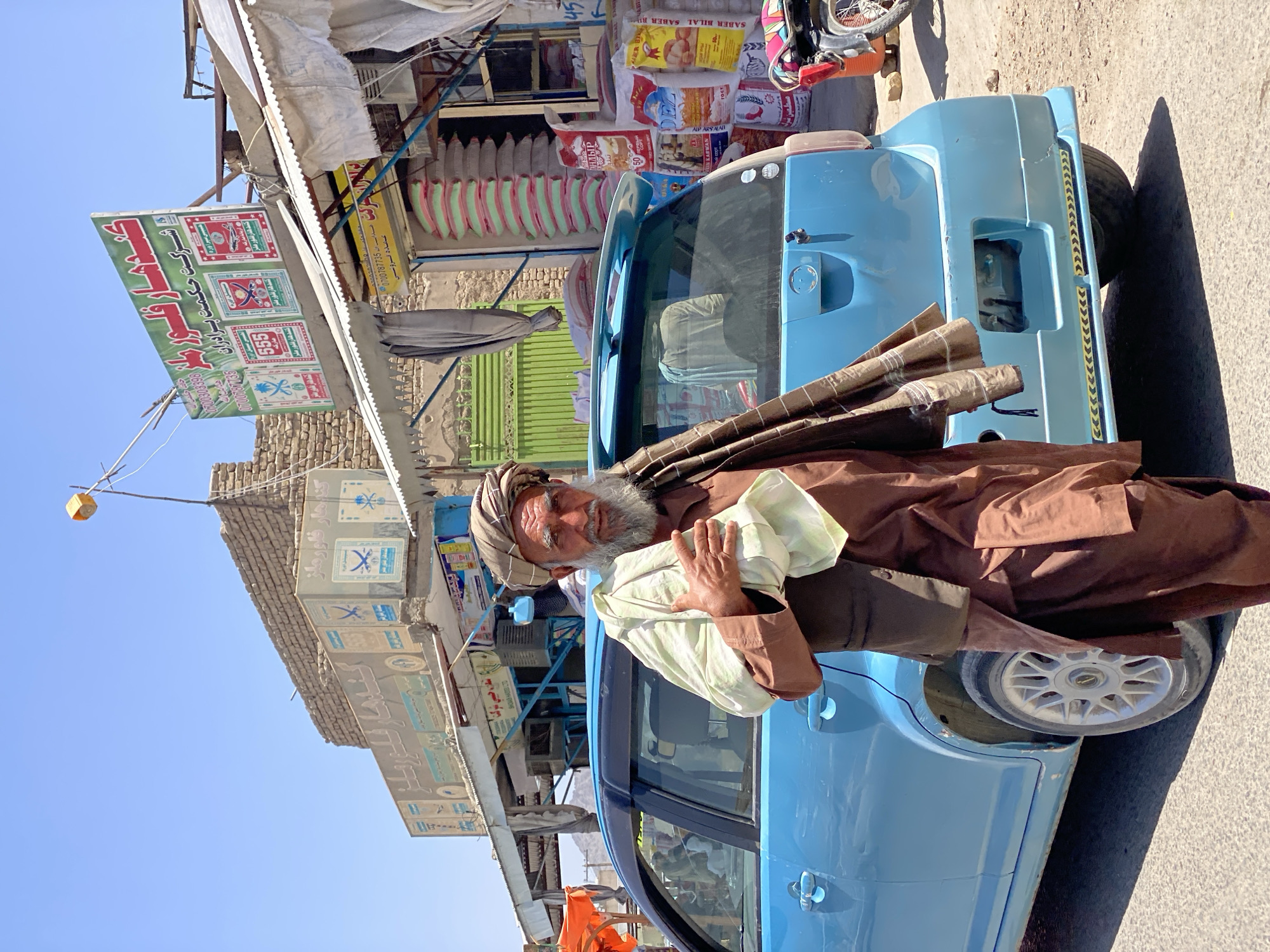 An outdoor, full-body, medium shot captures an elderly Afghan man with a grey beard and turban in the city of Kandahar, Afghanistan. He is standing in front of a bright blue car, holding a white cloth in his left hand. He is wearing a brown tunic and vest. Behind him, a store displays sacks of rice and other goods. The sky is clear and blue, and the scene is bathed in bright sunlight. The overall impression is one of a candid moment captured in a busy marketplace.
