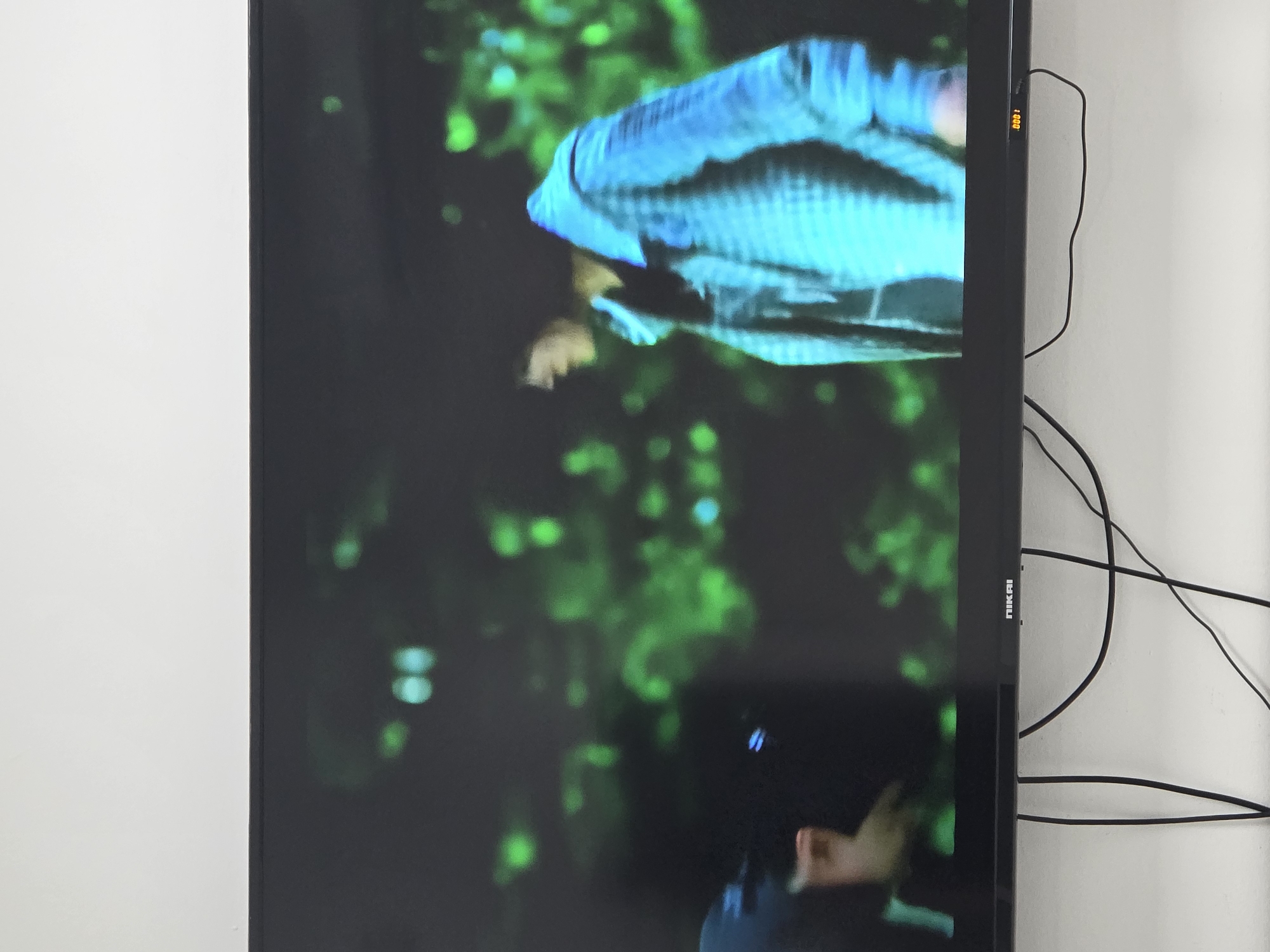 A wide, dark television screen displays a blurred image of two individuals against a background of out-of-focus green lights. On the left, a person wearing a dark helmet or hat is partially visible, with a hand extended. To the right, a person in a light blue, checkered jacket is shown in profile, looking downwards and to the left. The television itself is mounted on a white wall, with black cables hanging loosely below it. A logo reading "NIKAI" is visible on the bottom edge of the screen. The scene appears to be indoors, with the television screen being the primary focus. There is no discernible information within the image to determine the location in Dibba Province, Oman.