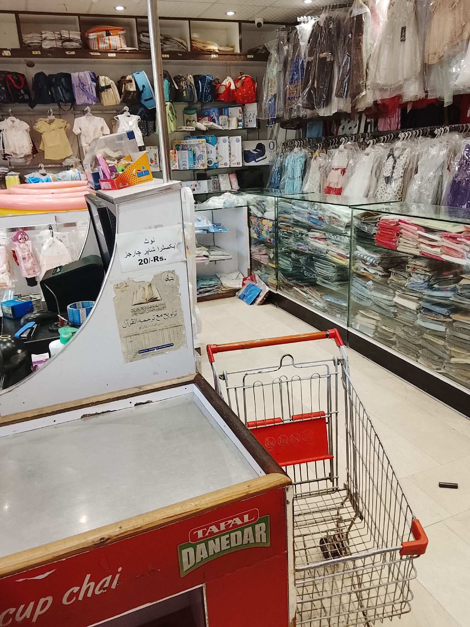 The image shows the interior of a retail store, likely a clothing or general merchandise shop. The location is Gulgasht Colony, Pakistan, as suggested by the text on the signs. The store has shelves stocked with various items, including clothing, baby products, and packaged goods. Clothes are seen hanging on racks, many in plastic coverings. Glass display cases are filled with neatly folded fabrics. At the forefront, there is a cash register with a sign in Urdu indicating an extra charge of 20 Rupees for shopping bags. A shopping cart stands nearby, suggesting customers are actively shopping. The environment appears well-lit and organized.
