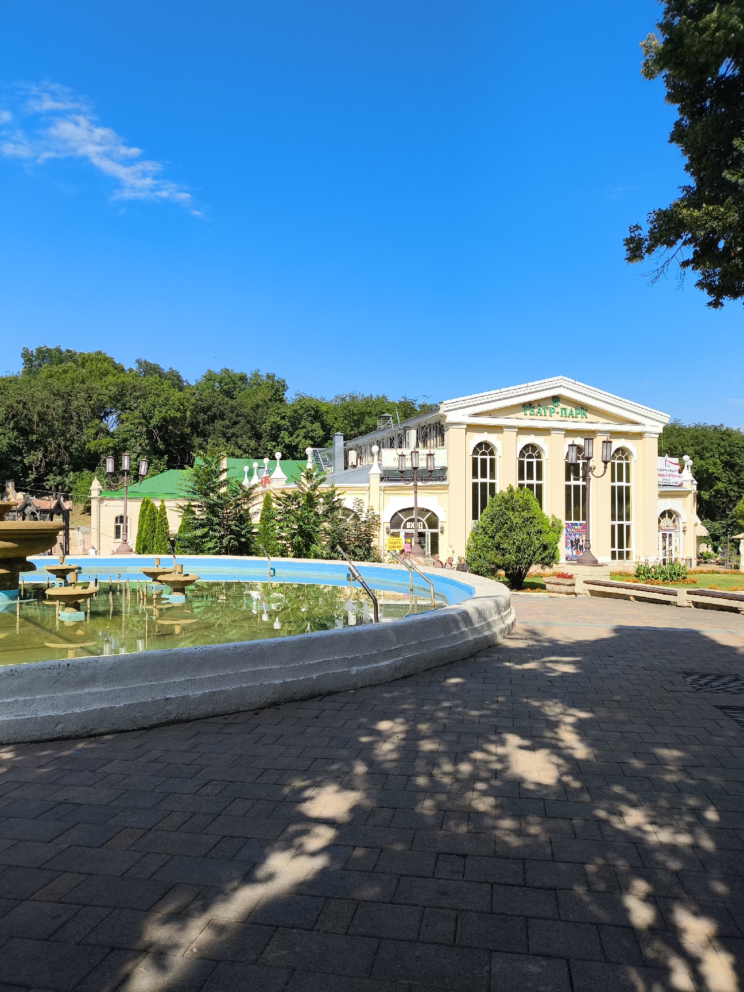 The image depicts a sunny day in Yessentuki, Russia, specifically in the vicinity of a theater. In the foreground, a large, ornate fountain with multiple tiers is visible, filled with greenish water. The fountain is situated beside a paved walkway made of dark gray bricks, with dappled shadows from trees falling across it. To the left of the fountain is a sidewalk and lush green trees.
Dominating the midground and background is a light yellow building with classical architectural features, including tall arched windows and a pedimented roof. The word "ТЕАТР-ПАРК" (Theater-Park) is inscribed in green letters on the facade. Several lampposts and smaller trees are present in front of the building, and people can be seen milling about on the grounds. Further behind the theater, a dense forest provides a backdrop, and a bright blue sky with a few wispy clouds completes the scene.