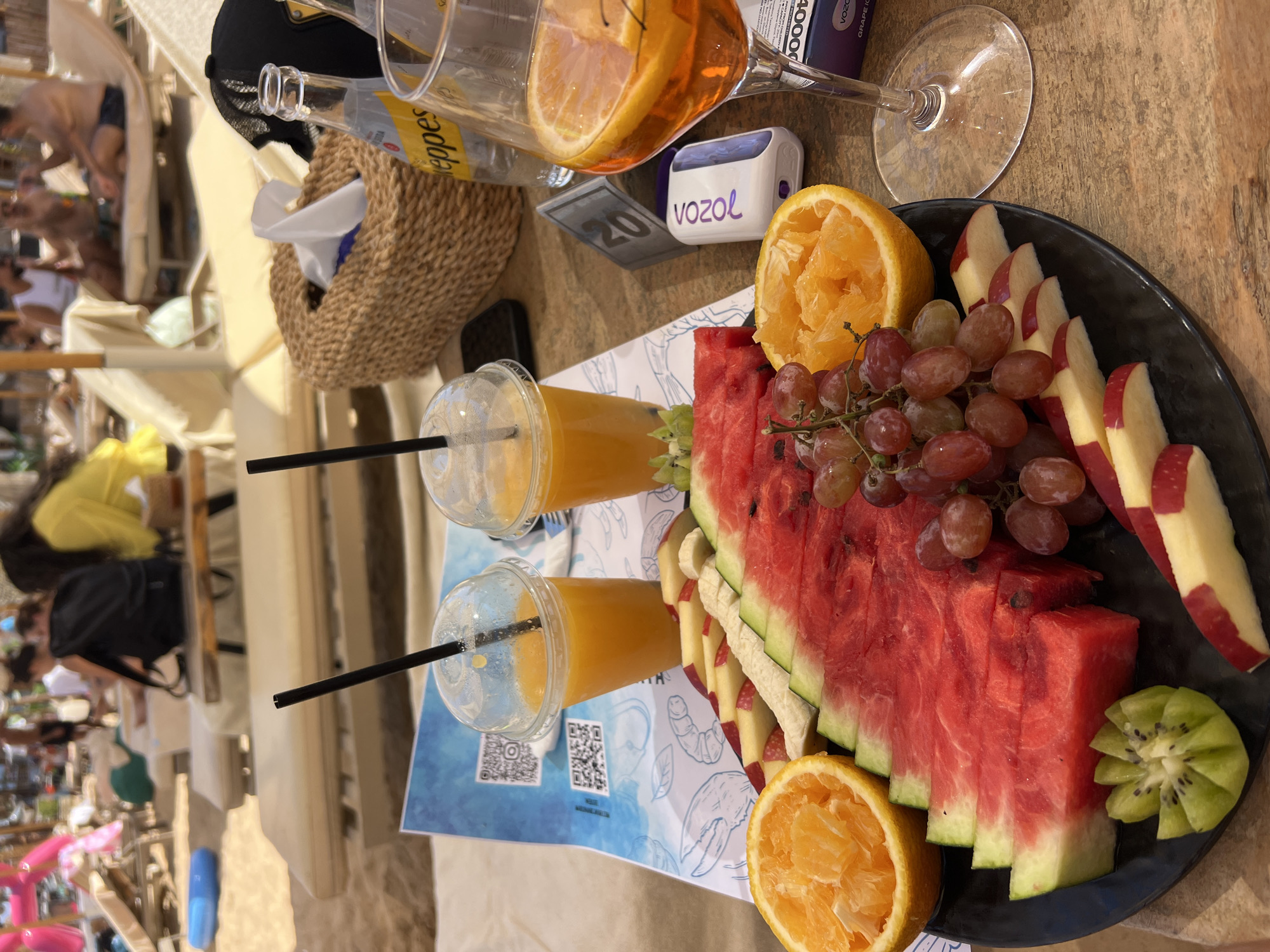The image depicts a beachside scene in Sharm El Sheikh, Egypt, with a focus on a platter of fresh fruit and drinks. In the foreground, a black plate holds a colorful assortment of fruits, including sliced watermelon, grapes, orange wedges, sliced apples, and a decorative kiwi slice. Two clear plastic cups filled with orange juice and black straws are positioned behind the fruit platter. To the right, a clear stemmed glass contains an orange-colored beverage with orange slices. A woven basket containing tissues and a bottle of Perrier water are also visible. In the background, beach chairs and umbrellas suggest a relaxing atmosphere, with blurred figures of people enjoying the sun. The wooden table surface and sandy beach backdrop further enhance the beach resort ambiance. The scene is brightly lit, consistent with an outdoor daytime setting.
