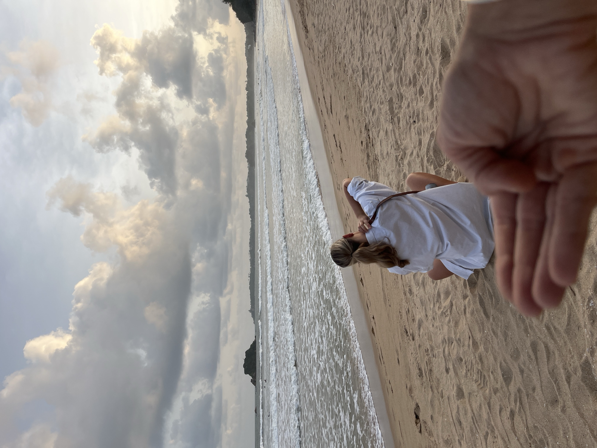 A person is sitting on a sandy beach, facing the ocean. The person is wearing a light-colored, striped shirt and has blonde hair pulled back. The ocean waves are gently rolling onto the shore. In the background, there is a distant shoreline with trees and a rocky outcrop. The sky is cloudy with patches of sunlight breaking through, suggesting it might be late afternoon or early morning. The overall mood appears peaceful and contemplative. The image is taken from a slightly elevated angle, with a hand in the foreground partially obscuring the view, indicating the photographer might be a person close to the subject. No text is visible in the image. The setting is Peleana, Sri Lanka.