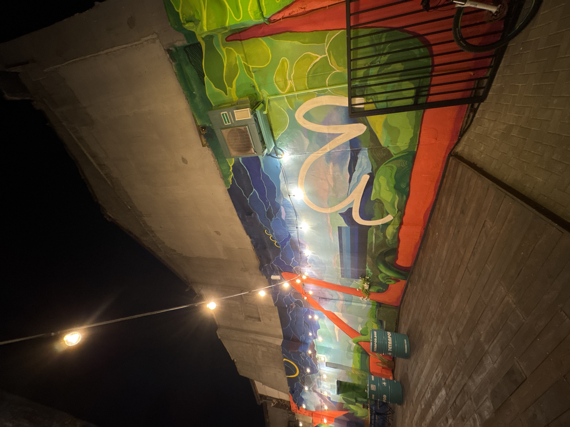 The image depicts a public space in Varna, Bulgaria, specifically underneath a bridge. The area features a mural painted on the bridge's supporting structures, depicting green foliage, a coastline with blue and white water, and stylized white shapes. An air conditioning unit is attached to the bridge support near the top of the mural. An orange structure with string lights is attached to the bridge and extends downwards. Several green barrels with the word "TETRAPOD" printed on them are positioned on the ground. The ground is paved with rectangular bricks. A black metal grating with a bicycle leaned against it is visible on the right. The scene is lit by the string lights, creating a nighttime atmosphere.