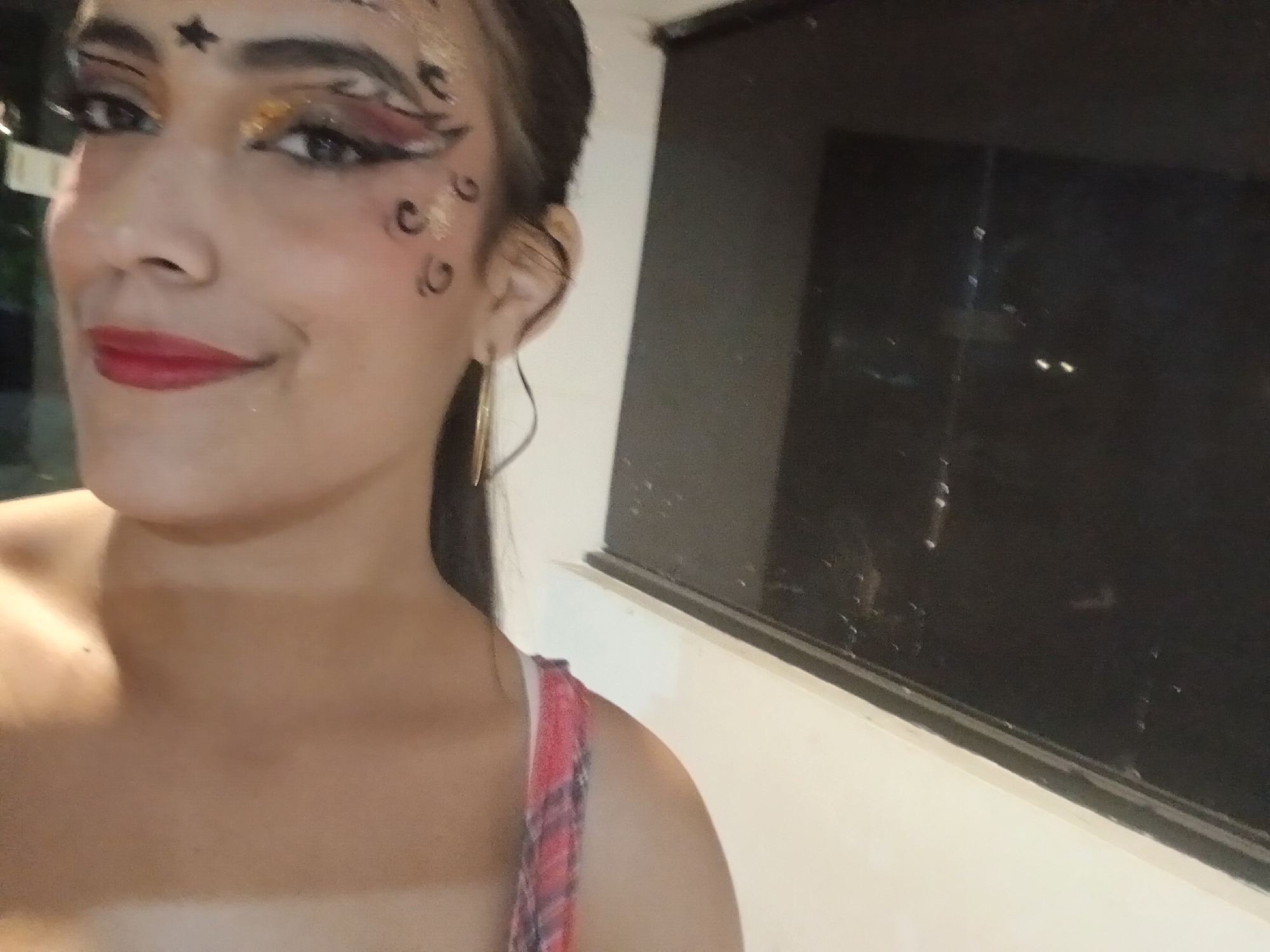 A close-up, slightly angled image captures a young adult female, visible from the chest up, looking directly at the camera with a slight smile. Her face features elaborate makeup, including a black star on her forehead, gold glitter eyeshadow, extended black eyeliner with red accents, and black curved markings with gold glitter on her left cheek. Her lips are adorned with red lipstick. She wears a gold hoop earring on her left ear and a red and white plaid strap on her right shoulder. The background is out of focus, revealing a dark, reflective window pane with faint light reflections and a white wall beneath it, indicating an outdoor evening or nighttime setting. The scene is located in Playa El Agua, Venezuela.