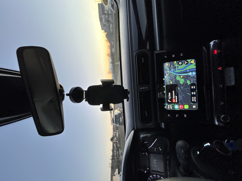 An interior view of a car, focusing on the dashboard and windshield. A rearview mirror hangs centrally, and a phone mount is attached to the windshield. A digital display shows a navigation map and driving information. A speedometer and other car controls are visible. The sky is visible through the windshield, with a gradient from blue to a warm sunset glow.