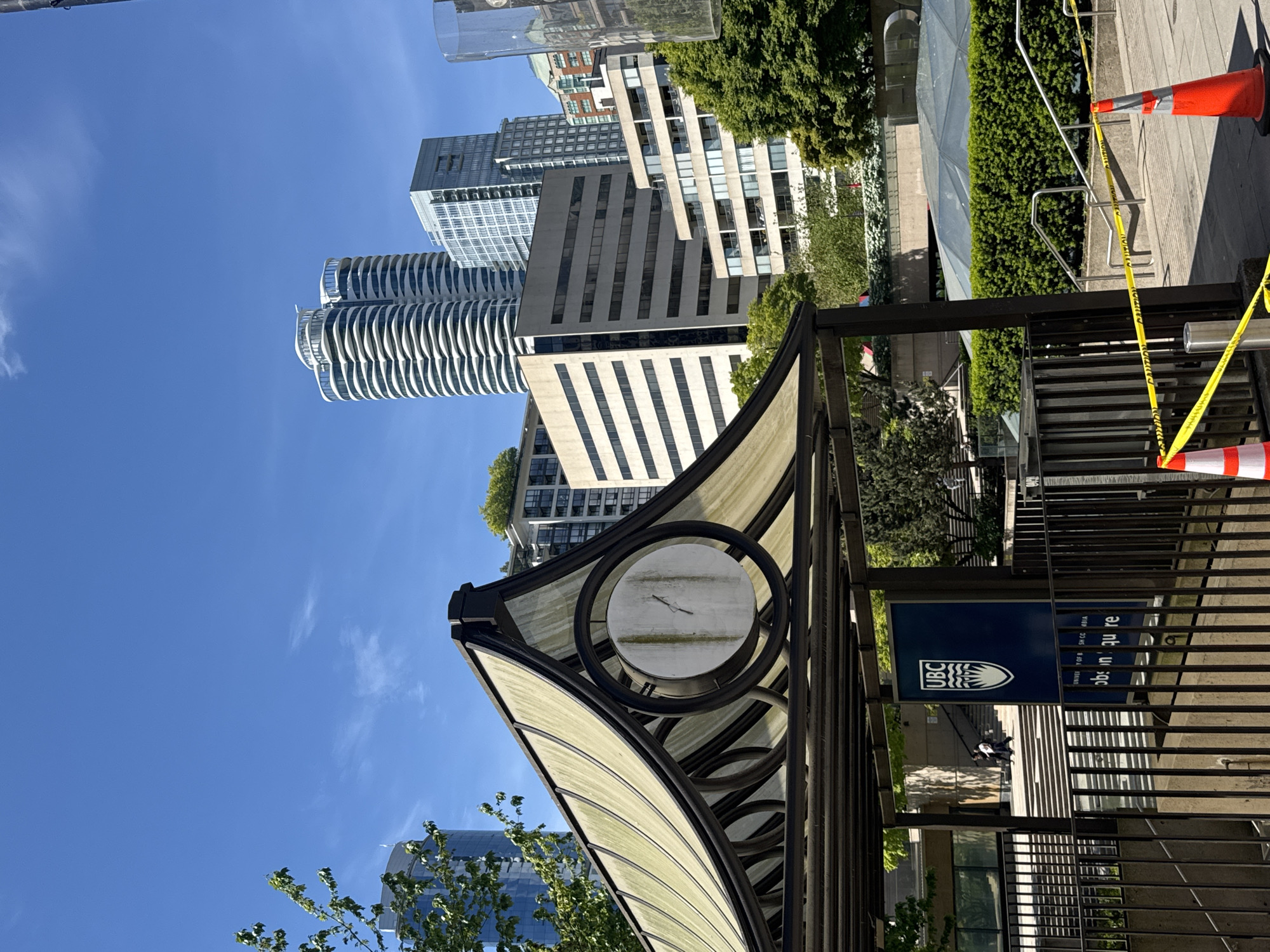 The image shows the UBC Robson Square campus in downtown Vancouver, Canada. In the foreground is the entrance structure with a clock and UBC signage. Buildings are visible in the background, and the sky is clear and blue. Construction tape and safety cones indicate that the area is closed or under repair. A glimpse of pedestrian traffic is visible in the background.
