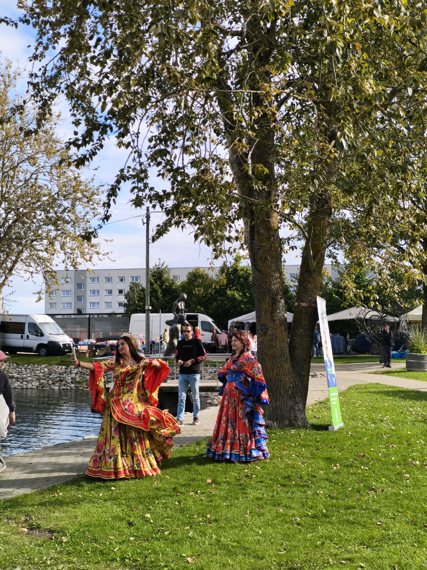 The image depicts an outdoor scene in the city of Maardu, Estonia, likely at a public event or festival. In the foreground, two women are dressed in elaborate, brightly colored, full-length skirts and tops, reminiscent of traditional folk costumes. The woman on the left, wearing a yellow and red outfit, is actively taking a selfie with her phone. The woman on the right is wearing a red and blue patterned dress. They are standing on a paved walkway next to a body of water, possibly a lake or canal.
Behind the women, several elements suggest a public gathering. There are market stalls with awnings visible in the background, and some people are browsing. A white van is parked to the left, and another is partially visible to the right. A statue or sculpture is also present behind the women, depicting figures in a dynamic pose. A large tree with green foliage dominates the right side of the frame, providing shade. The ground is covered in green grass, with some fallen leaves scattered about, indicating it might be autumn. The sky is partly cloudy, with patches of blue. A tall, light-colored apartment building is visible in the distance behind the event.