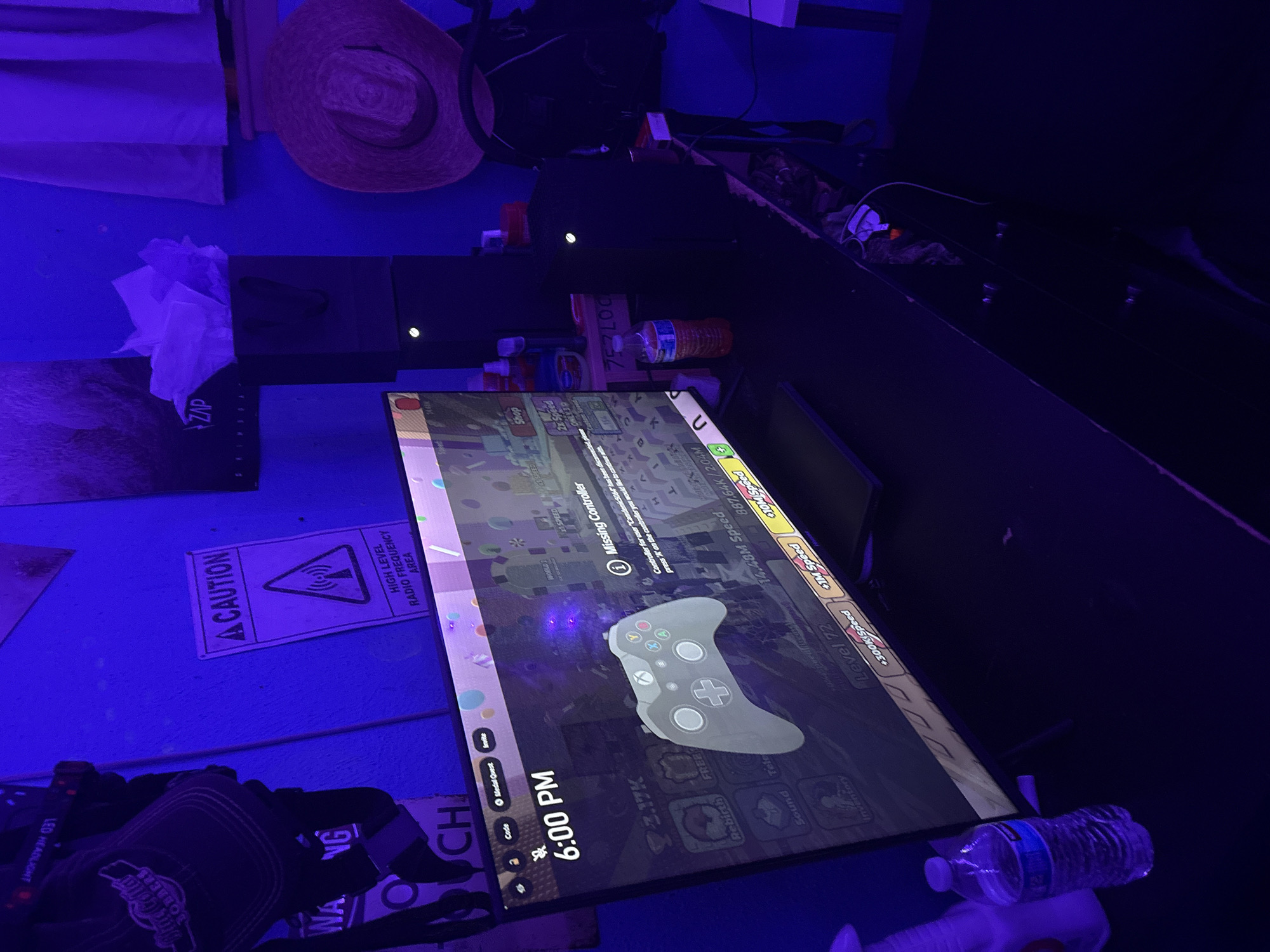 The image depicts a dimly lit room, possibly a bedroom or gaming den, illuminated by prominent blue and purple LED lighting. The primary focus is a computer monitor displaying a video game interface. The game shows a simulated Xbox controller and various in-game statistics and options, including "6:00 PM," "Missing Controller," and level progression information like "Level 72" and speed indicators.
On the desk next to the monitor are two black Xbox Series X consoles, stacked vertically. To the left of the monitor, a clear plastic water bottle and a white spray bottle are visible. On the wall behind the monitor, there is a "CAUTION" sign with a triangle and radio waves symbol, indicating a "HIGH LEVEL RADIO FREQUENCY AREA." Several other items are present in the room, including hats hanging on the wall, a straw hat on a shelf, and what appears to be a gift bag with tissue paper. The overall environment suggests a personal space dedicated to gaming, with a somewhat cluttered but organized setup. The image does not provide any specific geographical indicators to pinpoint a location within Virginia Beach, United States of America.