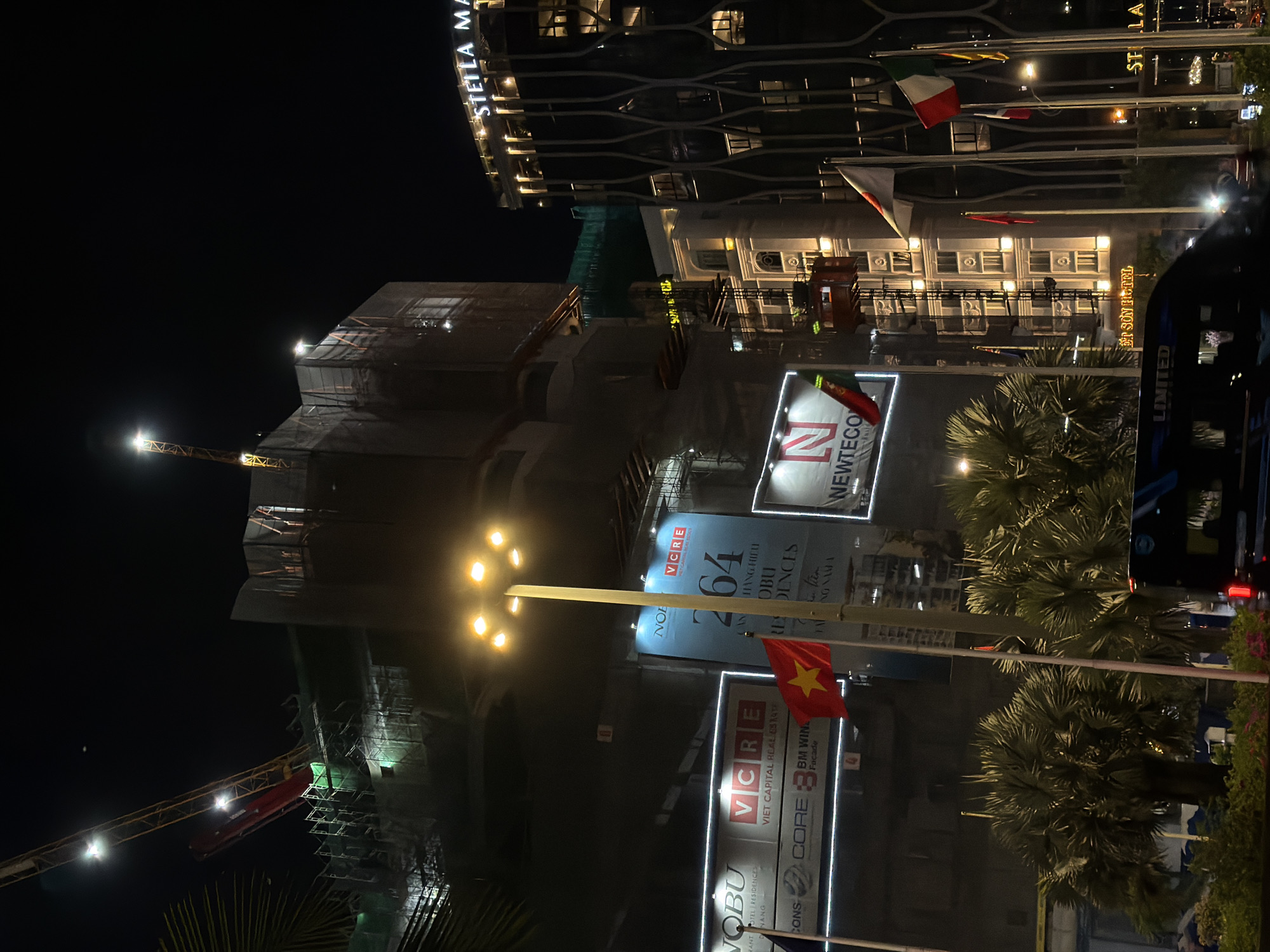 This is a night-time, low-angle shot of a construction site in Da Nang, Vietnam. The main subject is a tall building under construction, shrouded in scaffolding and netting. Several cranes are visible, including one yellow crane with lights and a red platform, and another red crane in the distance. In the foreground, palm trees are illuminated by artificial lights.
Behind the construction site, the façade of a hotel or luxury building is visible, with many lit windows. The text "STELLA MA" and "ET SON HOTEL" are visible on this building. Several flags are flying in front of the building, including the Vietnamese flag and Italian flag.
Large illuminated banners advertise the "VCRE" real estate company, and "NOBU HOTEL RESIDENCES". One banner prominently features the number "64" and text that appears to be Vietnamese. Another banner shows the text "NEWTECO". The overall impression is one of urban development and ongoing construction at night, with a backdrop of established, lit buildings and palm trees. The lighting suggests it is a clear night.